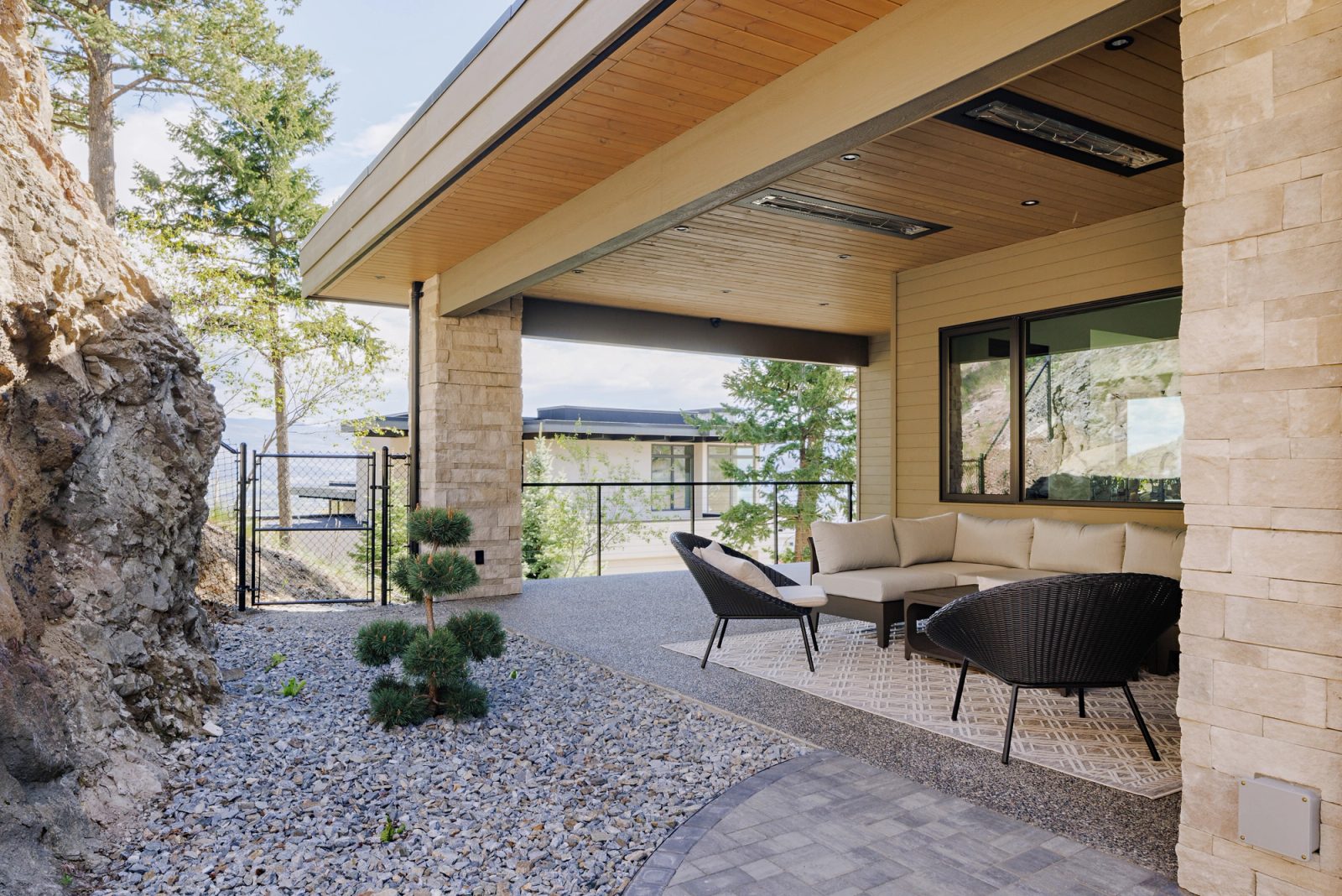 A covered outdoor room floored in exposed aggregate, wicker seating under a board soffit with radiant heaters, the rock face rising a metre from the furniture.