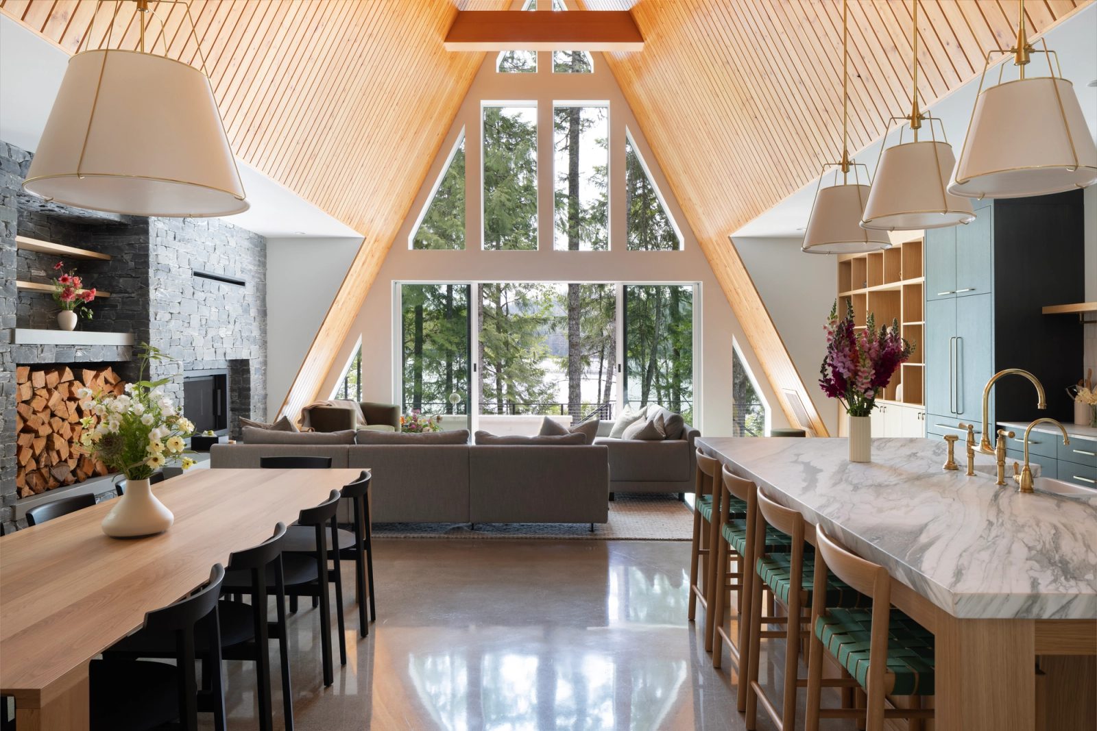 Great room under a pale cedar cathedral ceiling, stone fireplace at left, glazed gable open to the lake