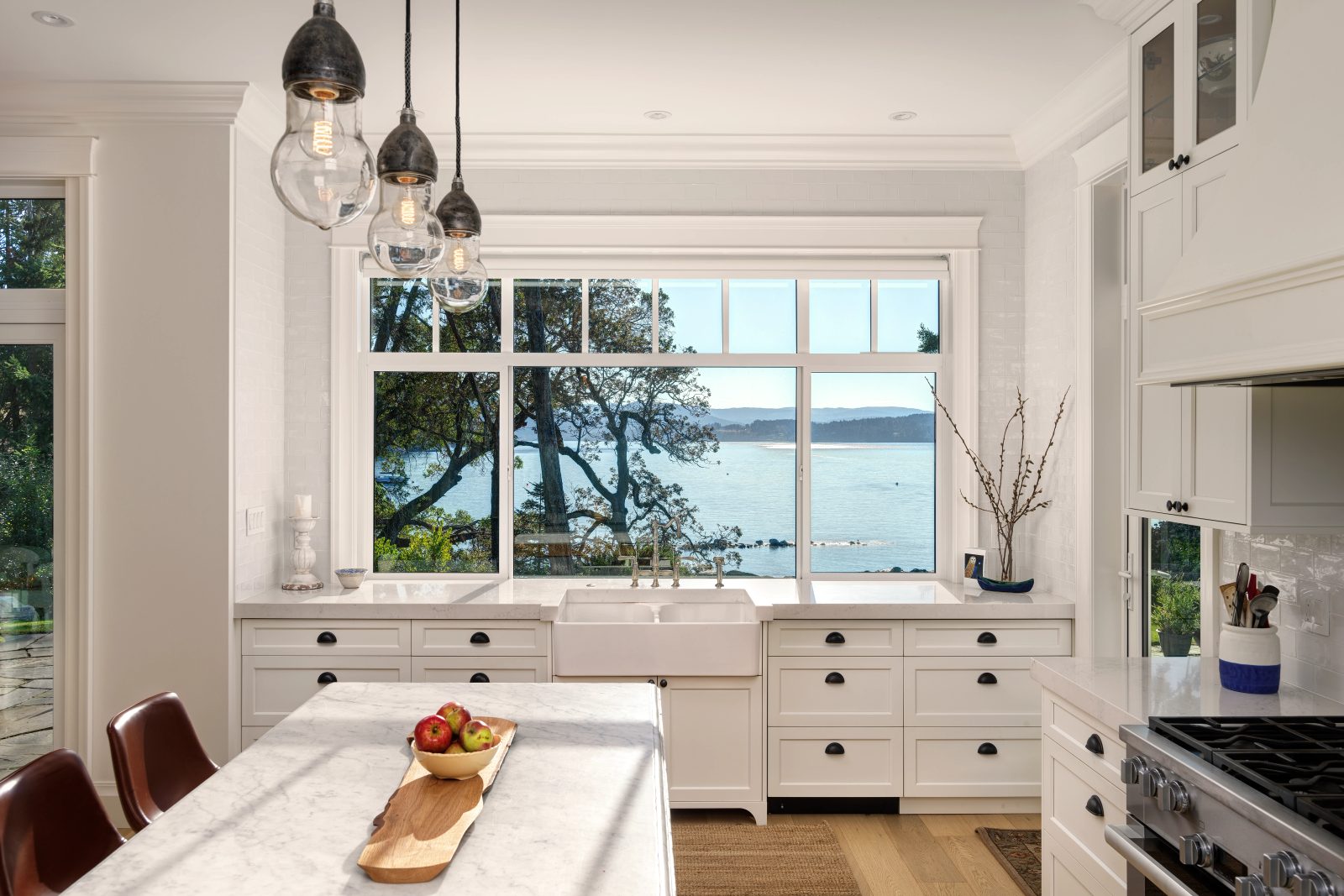 Window above an apron-front sink looking through arbutus branches to a bay and low hills across the water.