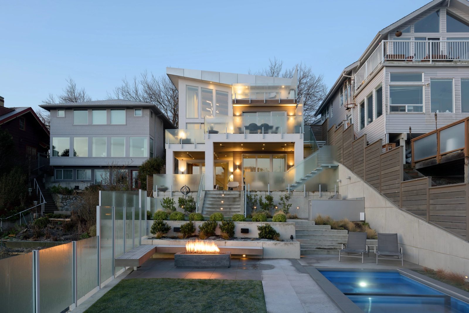 Three stepped storeys lit from within at dusk, seen across a lawn and a lap pool with a fire trough burning alongside.