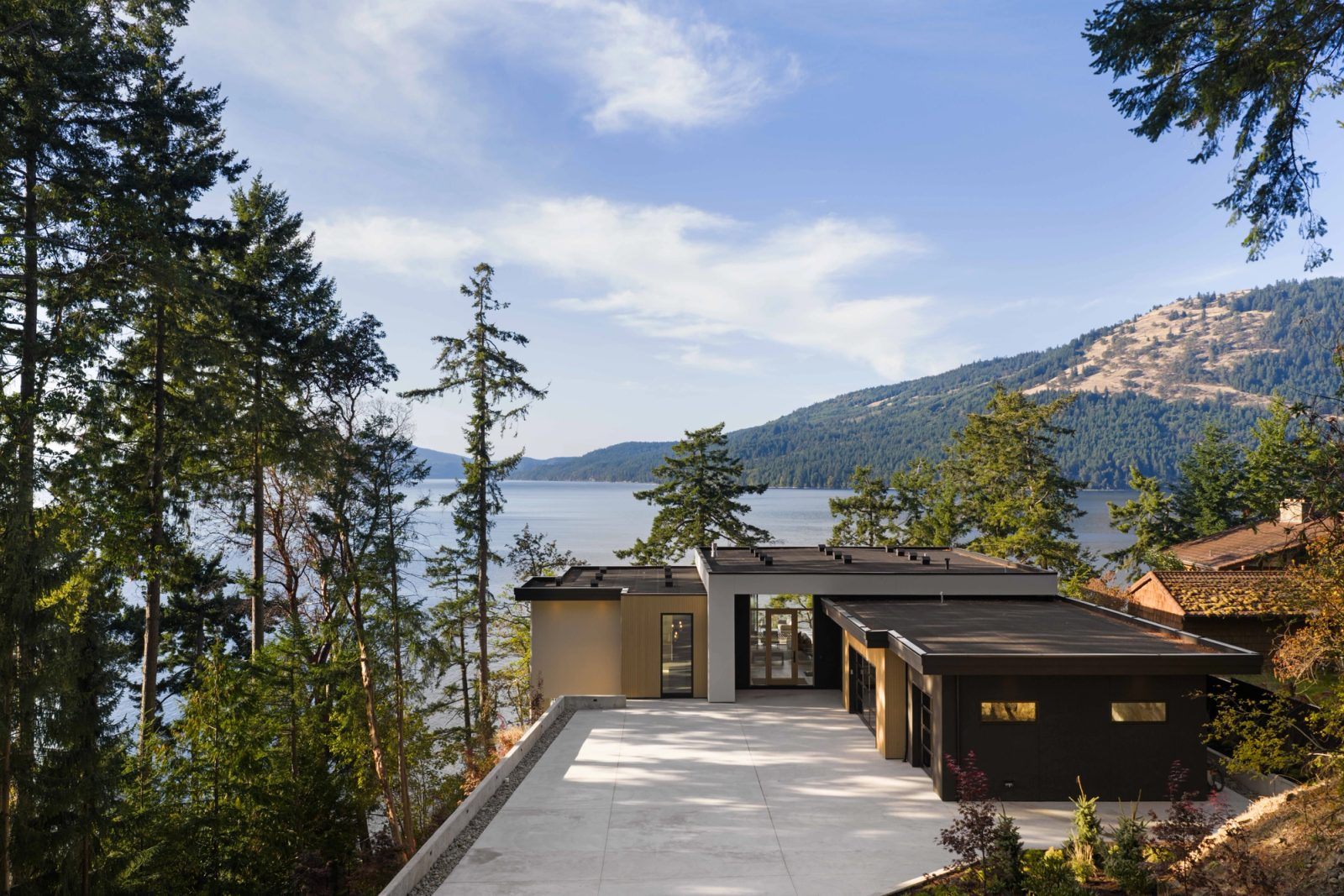A low flat-roofed house on a wooded point seen from above, its concrete forecourt running to the entry, open water and a forested ridge beyond.