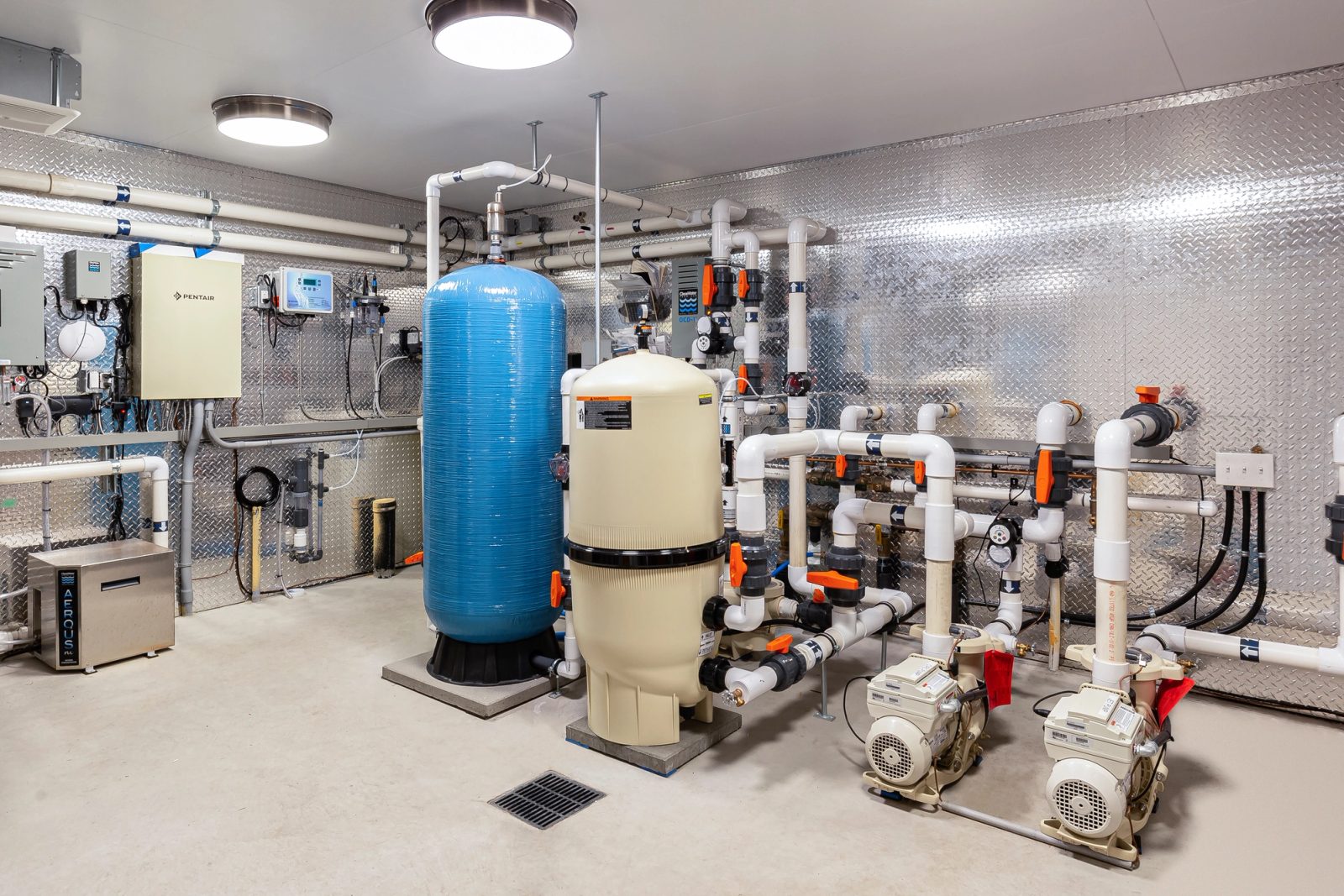 Pool plant room with filter tanks, pumps and valved white pipework against diamond-plate walls