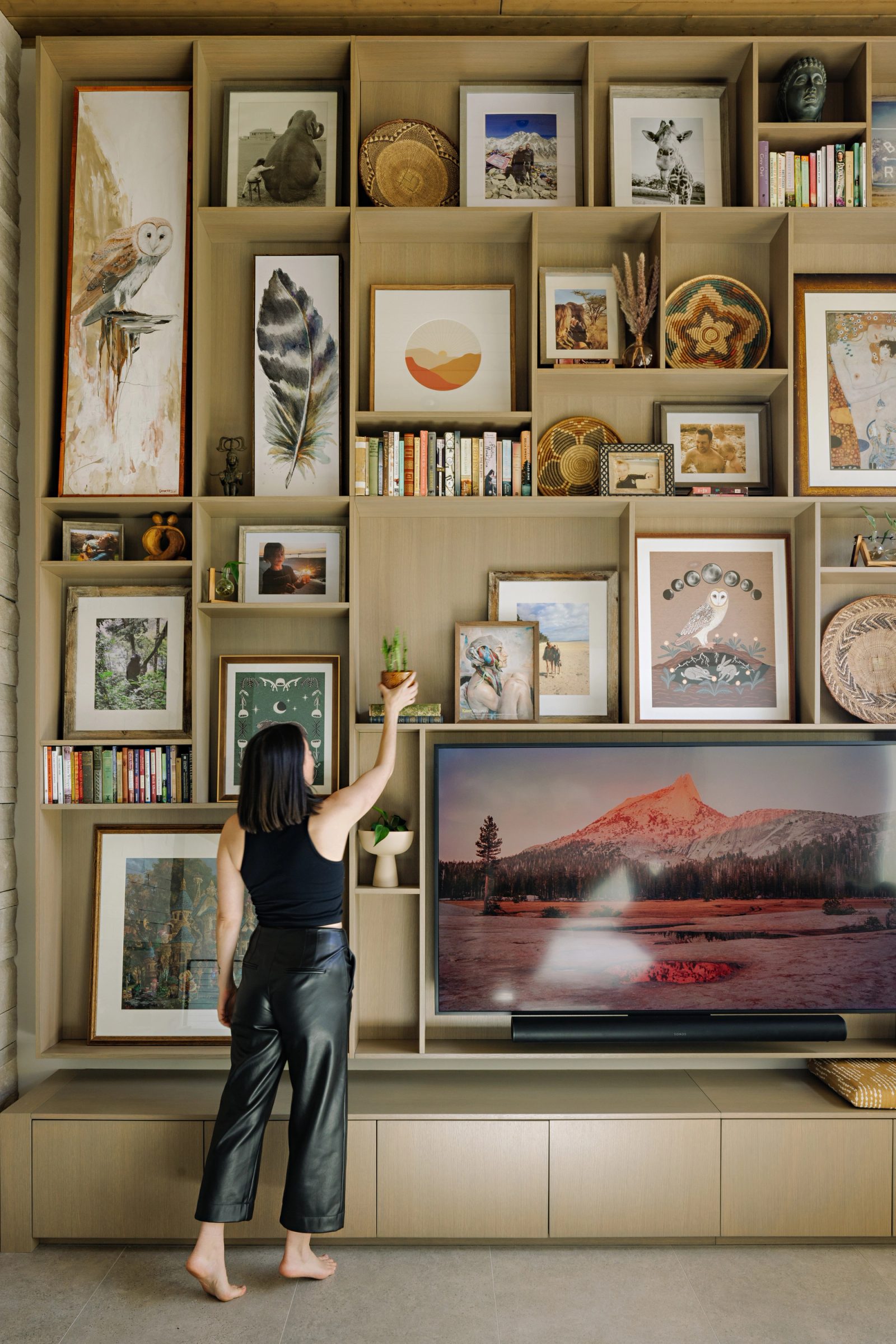 a floor-to-ceiling grid of pale wood shelving filled with framed pictures, woven baskets and books, with a figure reaching to place a small plant on an upper shelf.