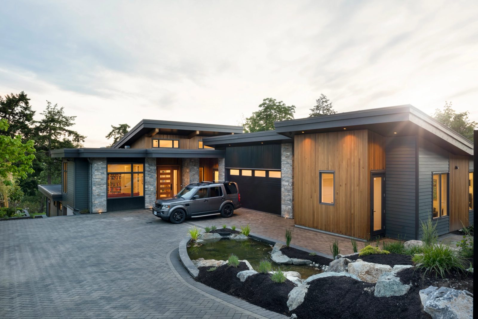 Single-storey modern house with cedar cladding, dark siding and stone piers above a paver driveway and planted pond