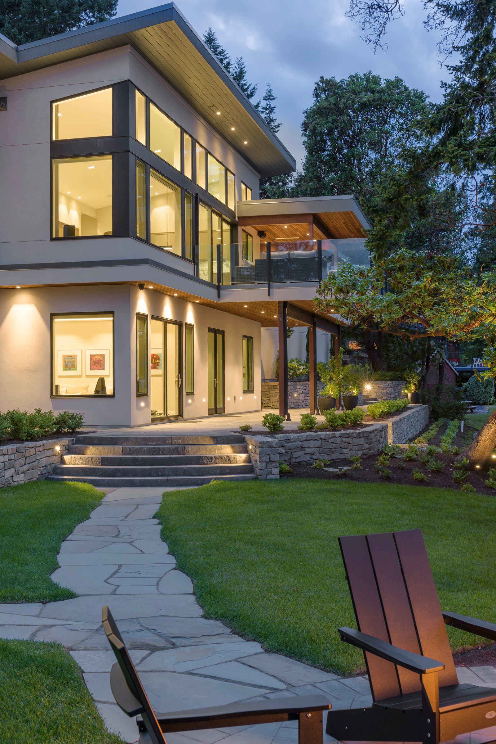 Waterfront elevation at dusk with a cantilevered deck, lit interiors and stone steps down to the lawn