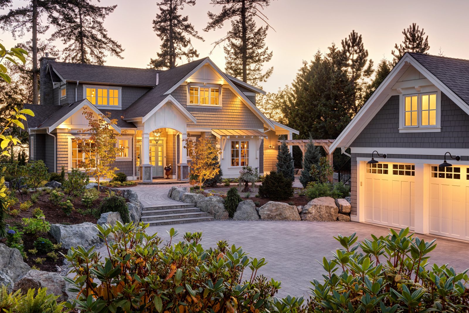 House and detached garage at dusk seen from the paver motor court, with rock outcrops and planting in the foreground and firs behind.