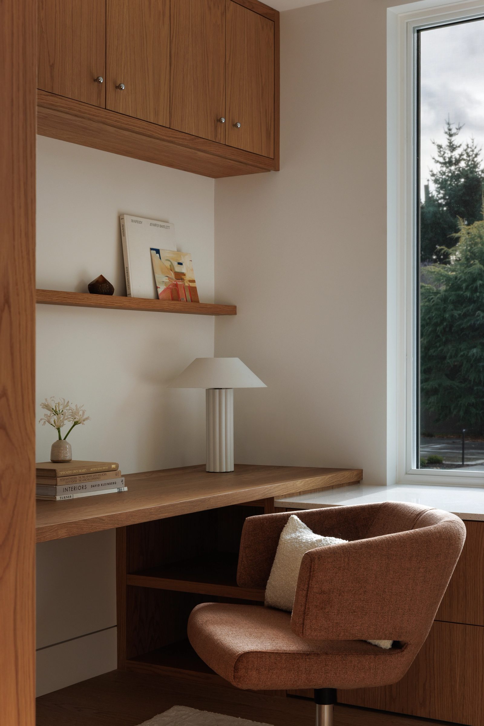 Left: a desk nook built in oak beneath cabinets and a floating shelf, a swivel chair drawn up to it and firs at the window.