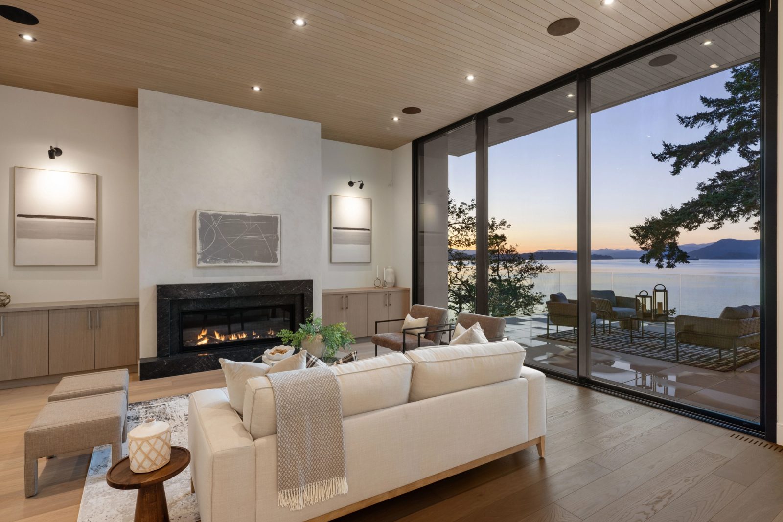 The same living room after sunset, the fire lit and the terrace lamps on, the last colour going out of the sky over the water.