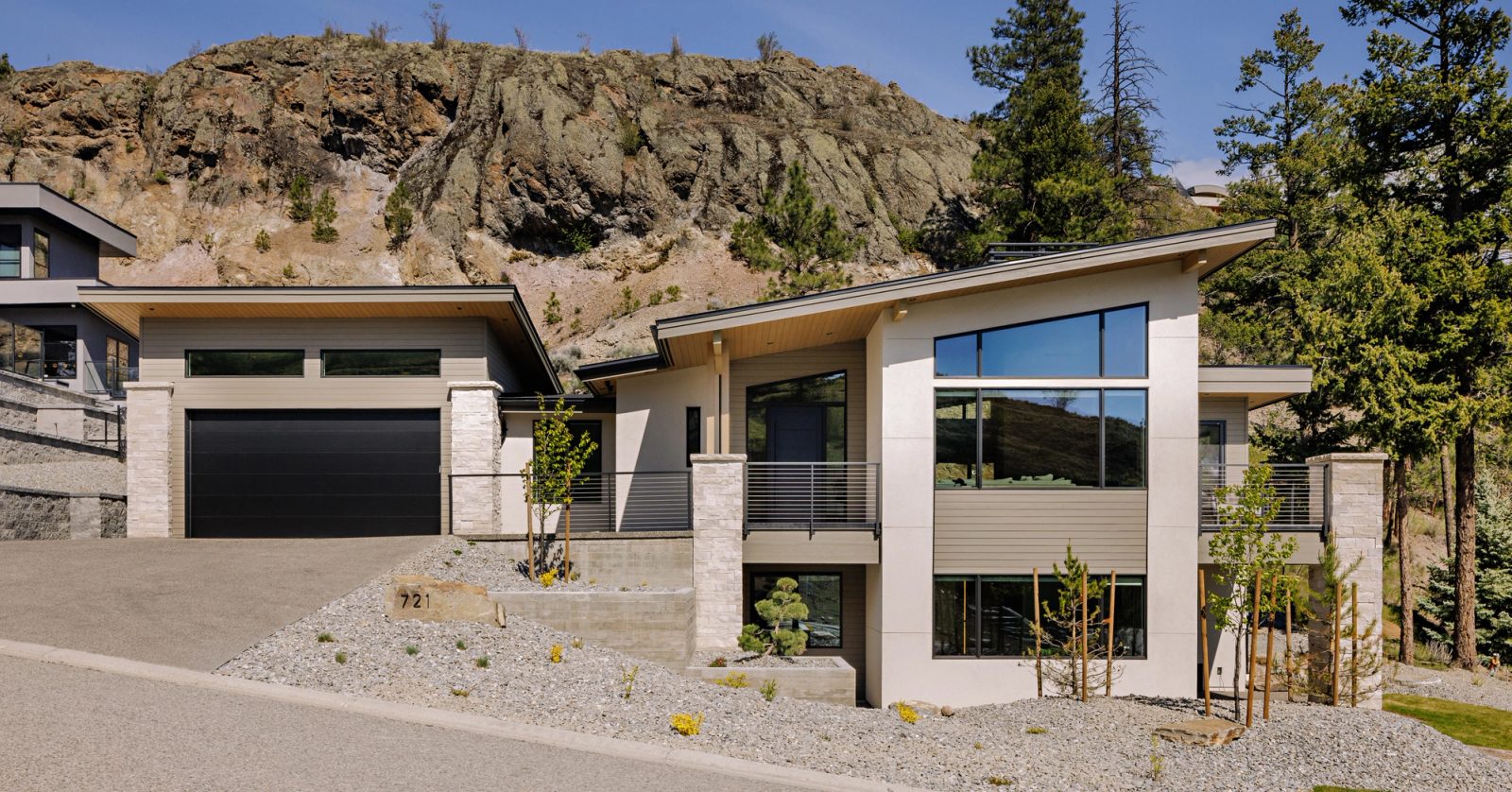 A house on a hillside lot below a bare rock bluff, with a double garage on the left and a two-storey window wall on the right, pines rising behind.