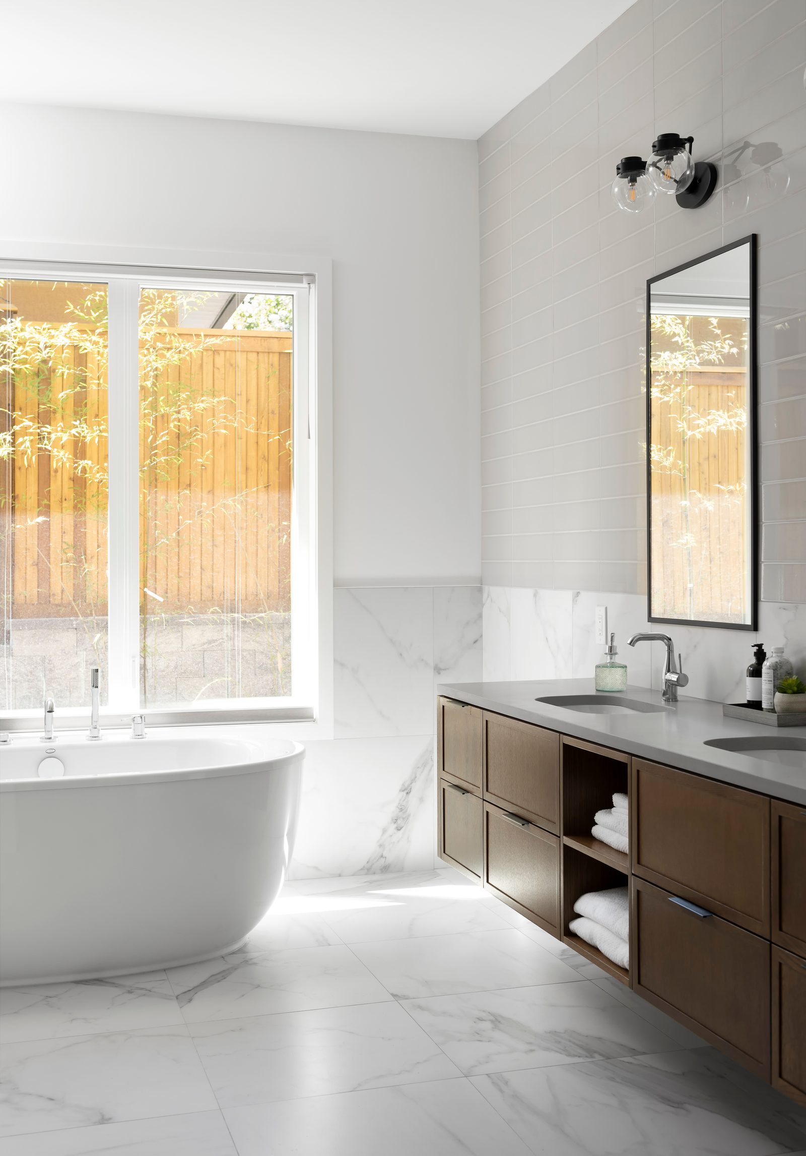 Ensuite with a freestanding tub under a window, marble-look tile and a double oak vanity