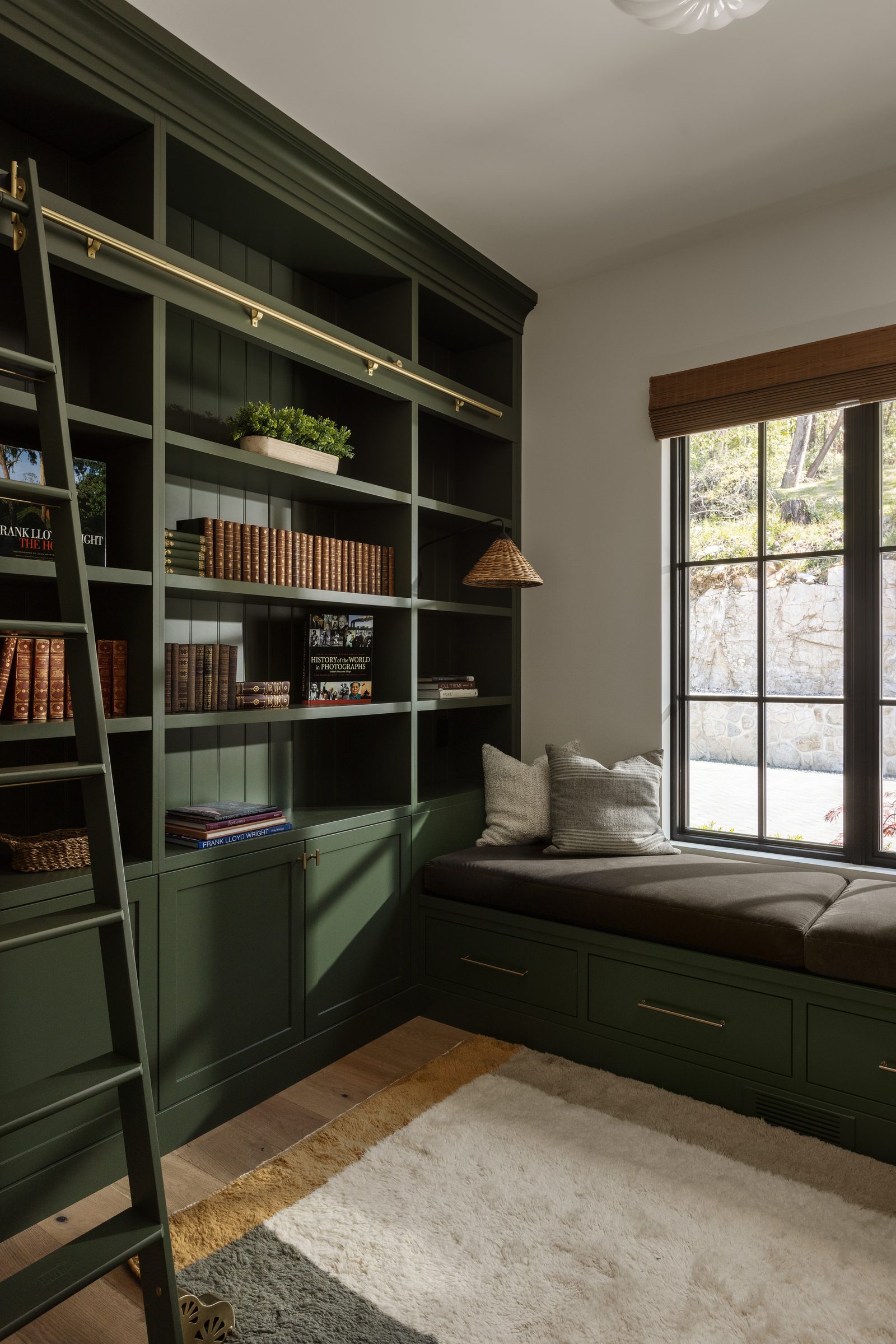 Right: a study lined floor to ceiling in dark green cabinetry with a brass library ladder, a cushioned window seat under a black-framed window.