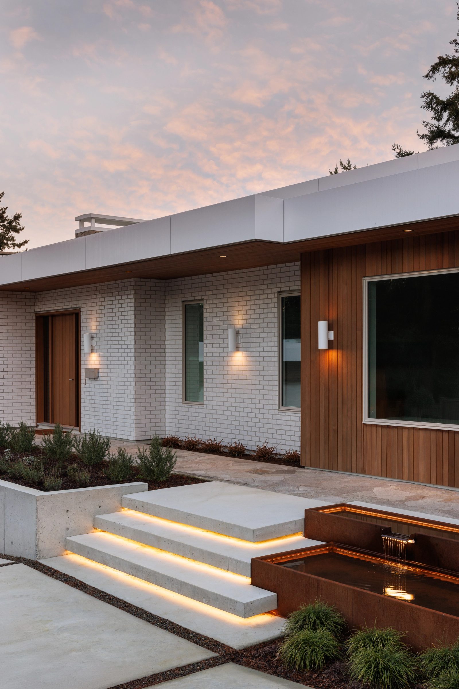 Left: the entry at dusk, sconces lit along white brick, concrete treads lit from beneath and a weathered steel basin alongside.