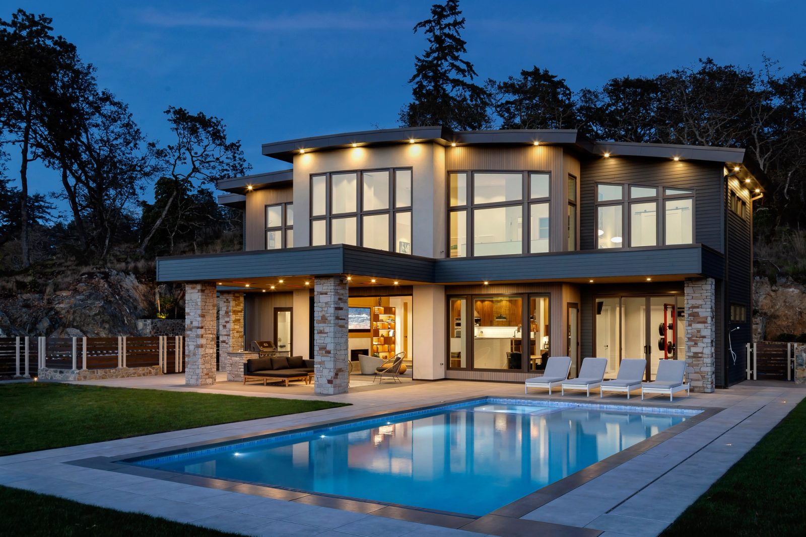 Lit modern house at dusk reflected in a rectangular pool, forested rock bluff behind