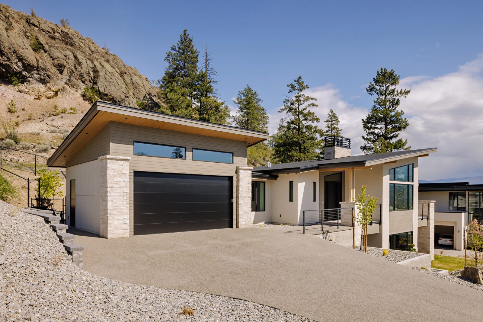 A wide concrete driveway running to a dark garage door set between stacked stone piers, with a cut rock face rising immediately behind the building.