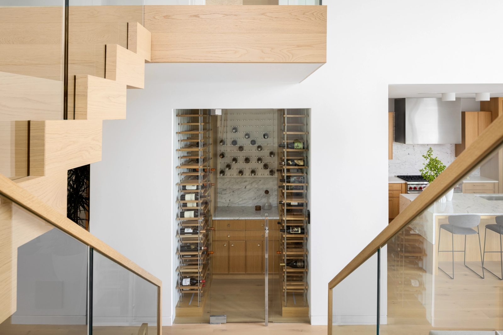 Glazed wine room seen between an open-tread stair and a glass guard, bottles racked on steel posts to either side.