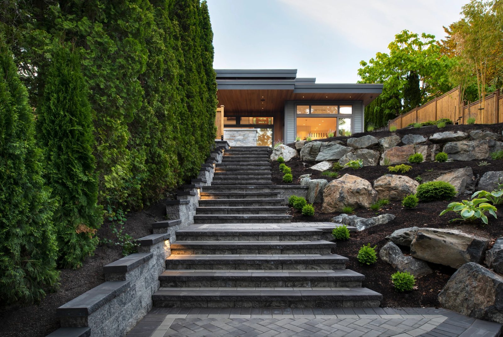Lit stone steps climbing between a cedar hedge and a boulder rockery to a house with deep cedar soffits