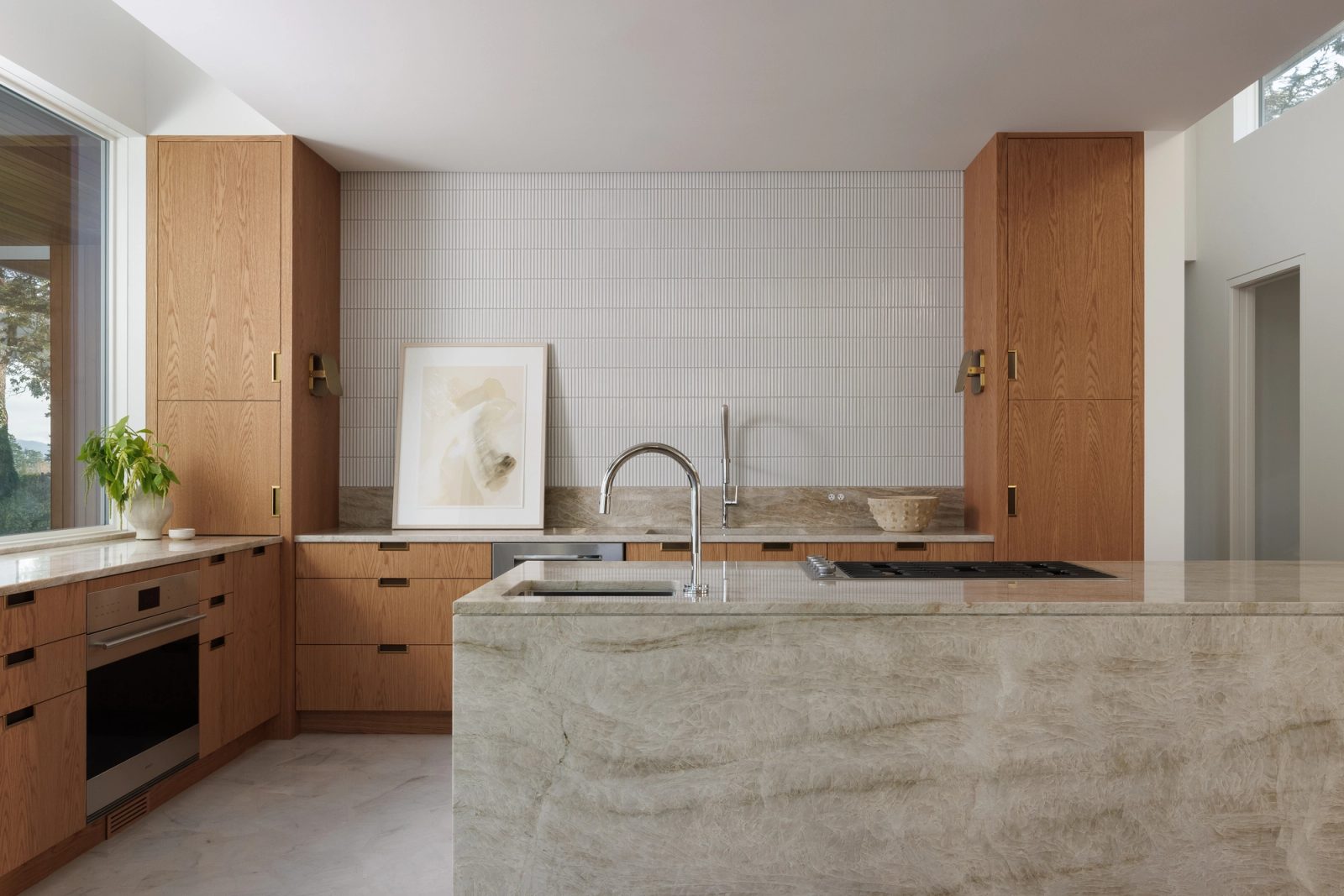 A kitchen with a quartzite island under a white fluted tile wall, oak cabinetry to either side and brass sconces on the returns.