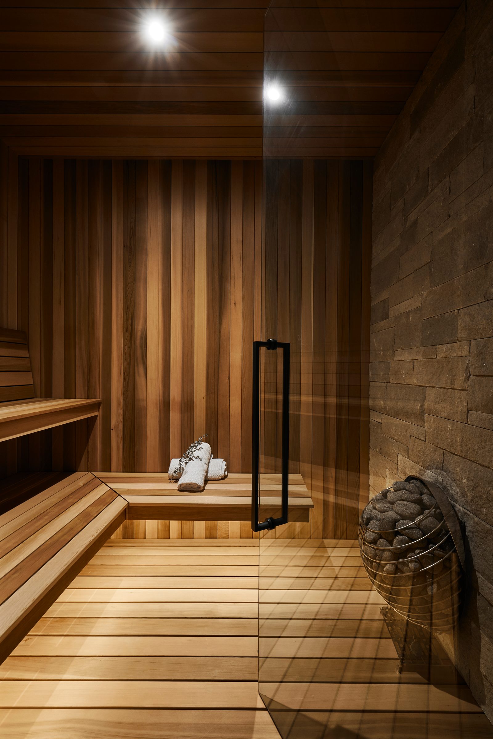 Cedar sauna with tiered benches, stone wall and heater behind a glass door