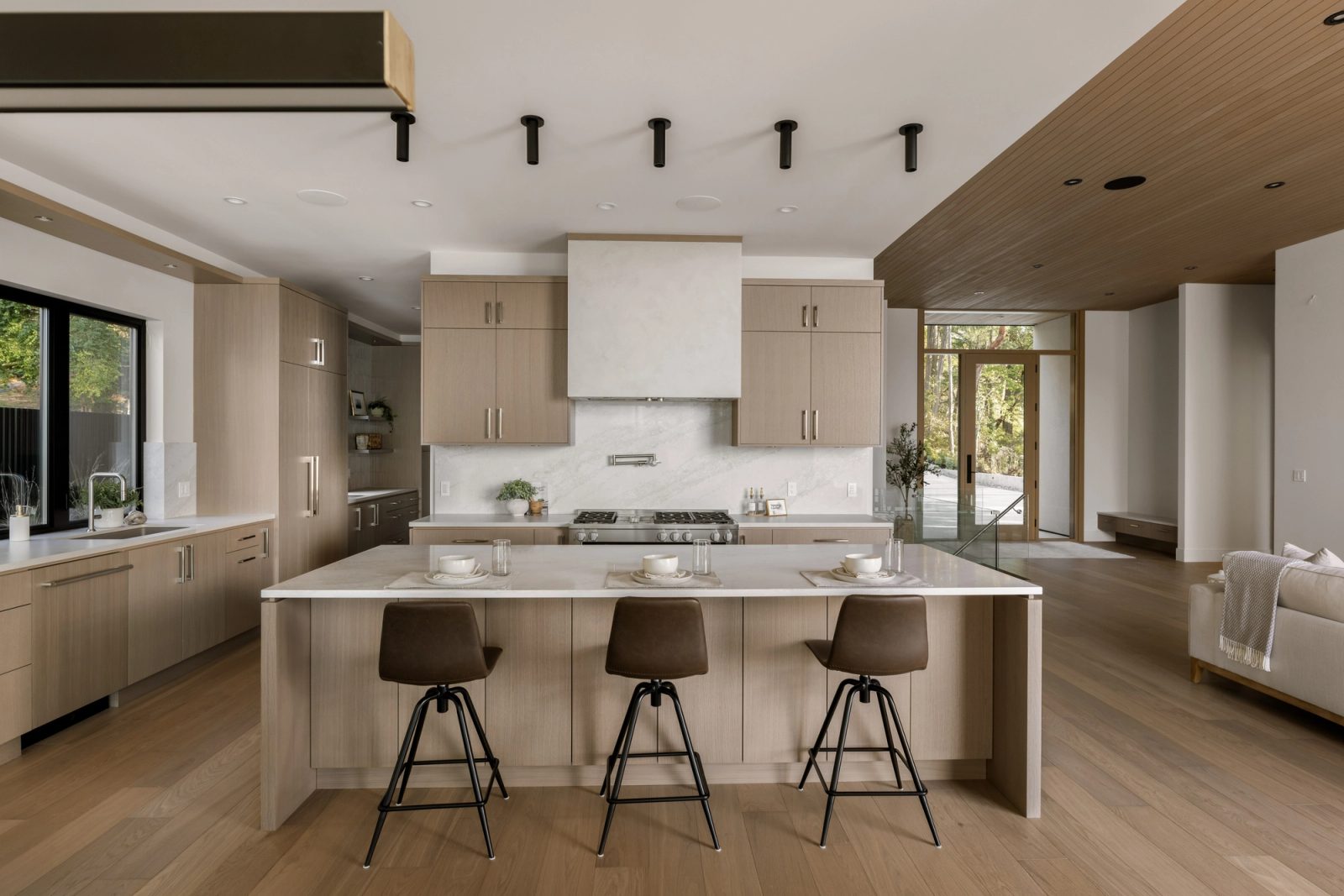 A kitchen in pale rift oak with a stone island and three leather stools, looking through to an entry glazed on both sides.