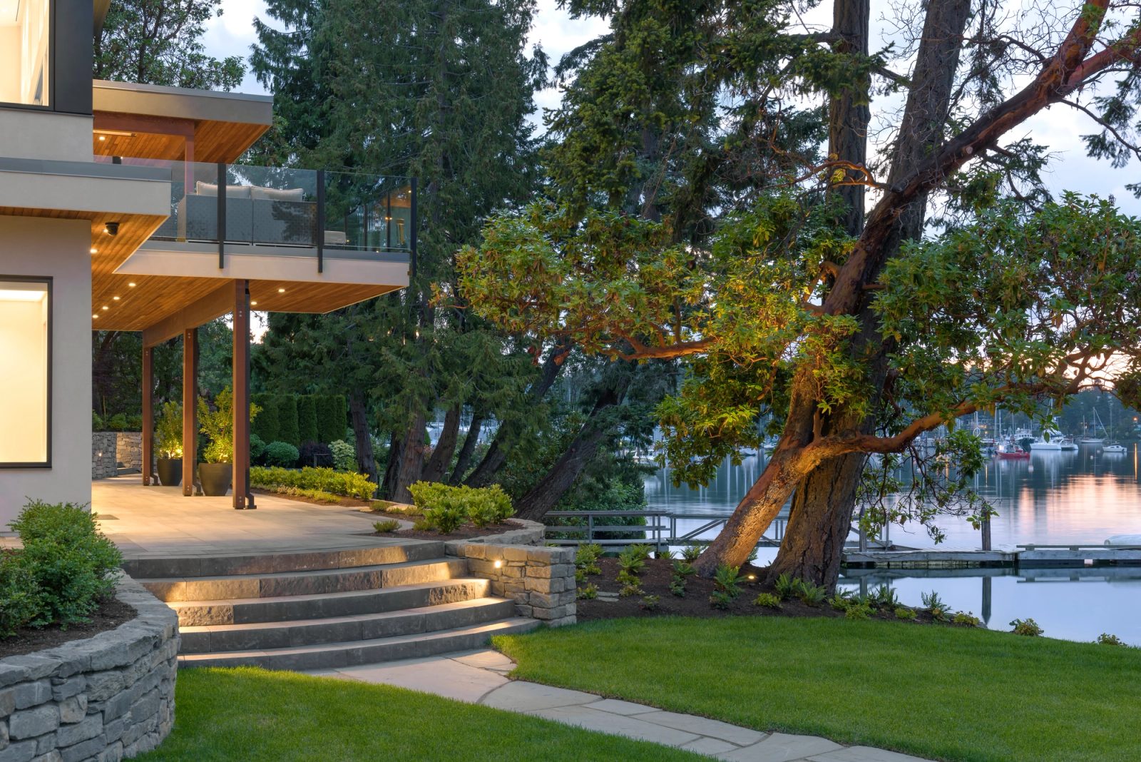 Cantilevered deck above a stone terrace, with an arbutus leaning over the lawn and moored boats beyond