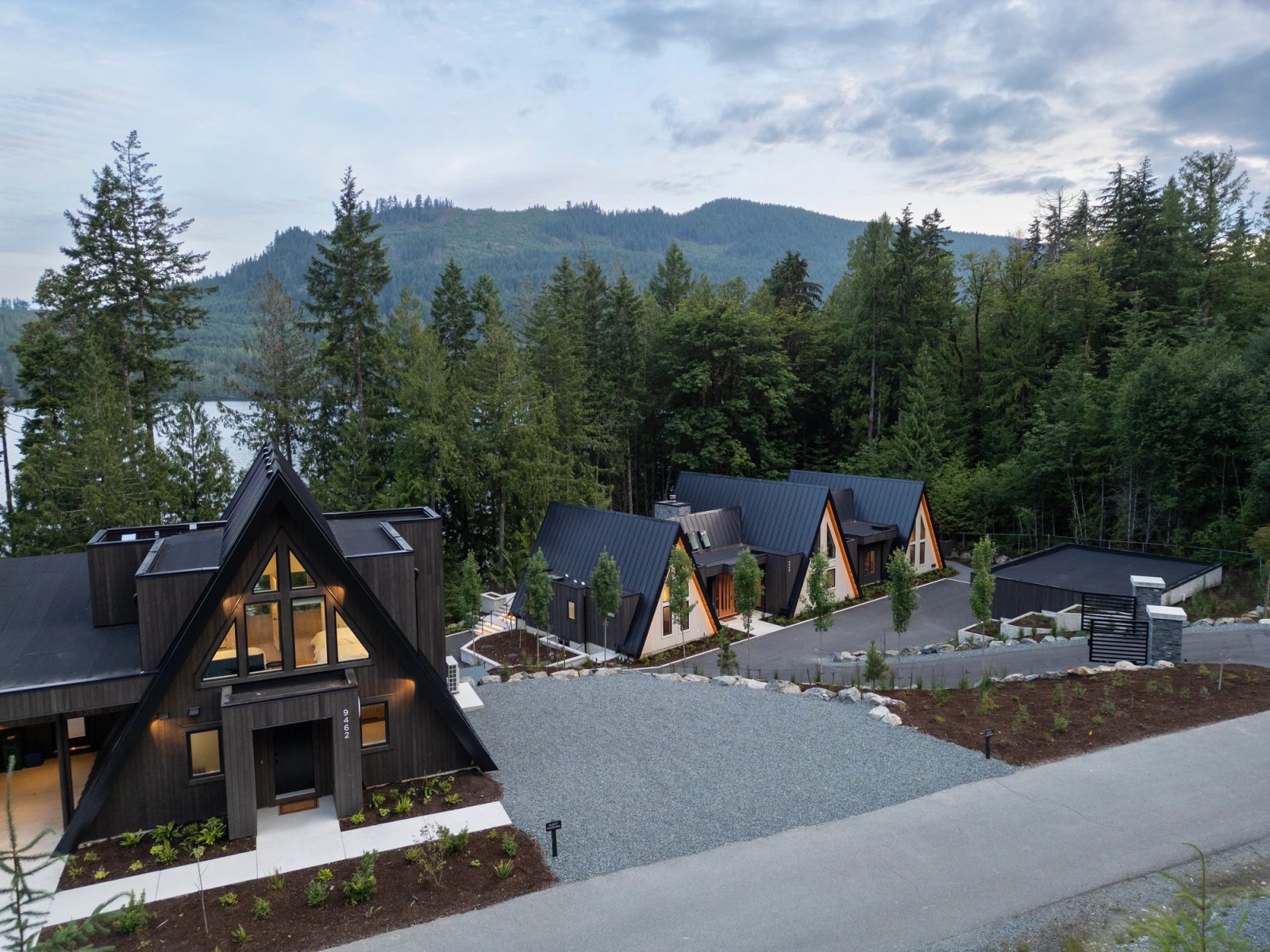 Aerial view of four black-clad A-frame buildings angled across a forested lakefront slope at Cowichan Lake