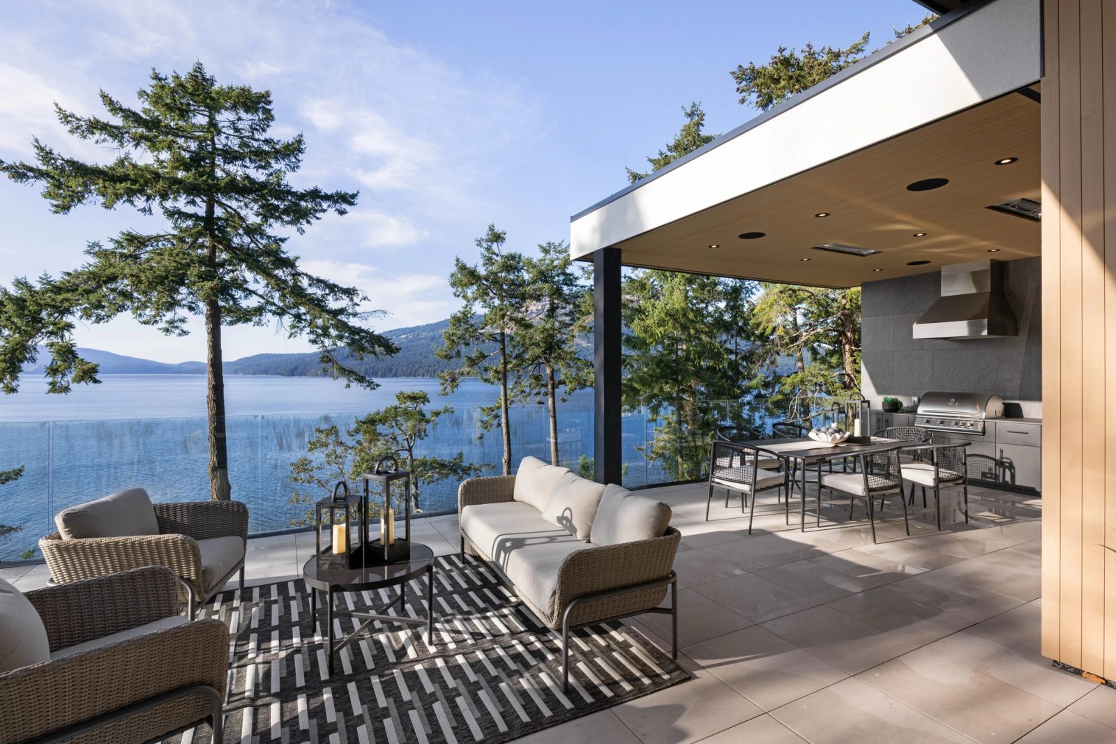 A covered terrace floored in large pale tile, woven seating and a dining table under a board soffit, glass rail and standing firs between it and the channel.
