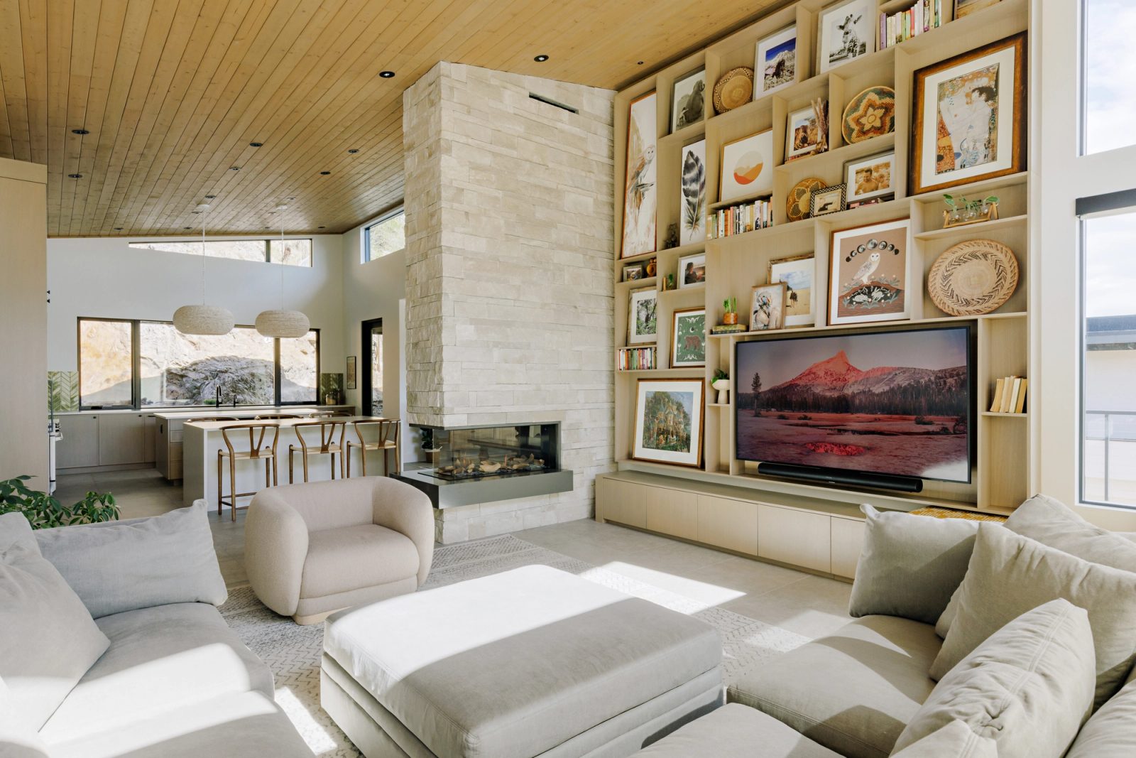 An open living room under a board-lined vaulted ceiling, a stone chimney standing free at the centre with a glass-sided firebox, and a kitchen visible beyond.