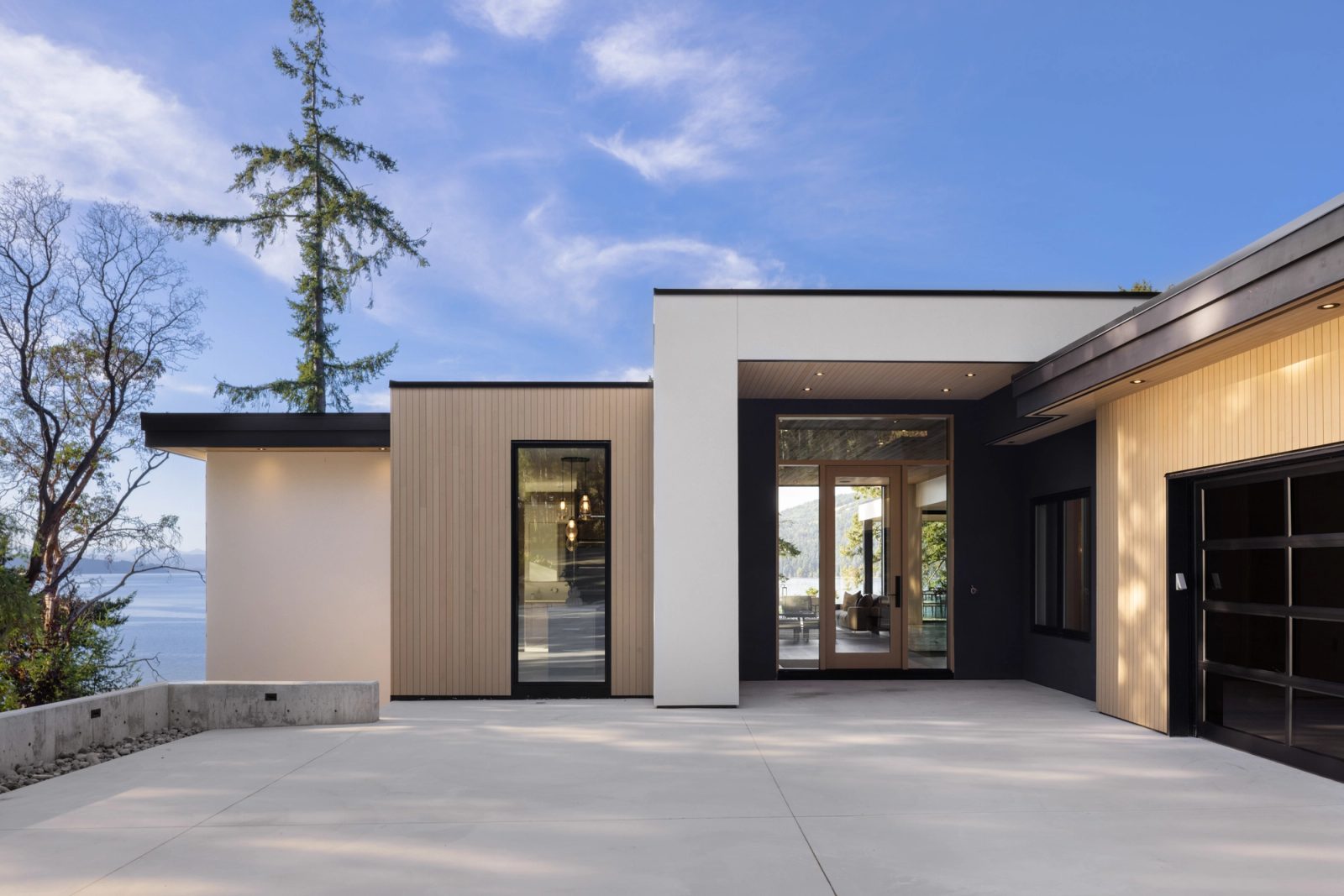 A wide concrete forecourt facing a recessed entry between pale vertical cedar and a dark stucco garage wall, the channel visible past the corner of the house.