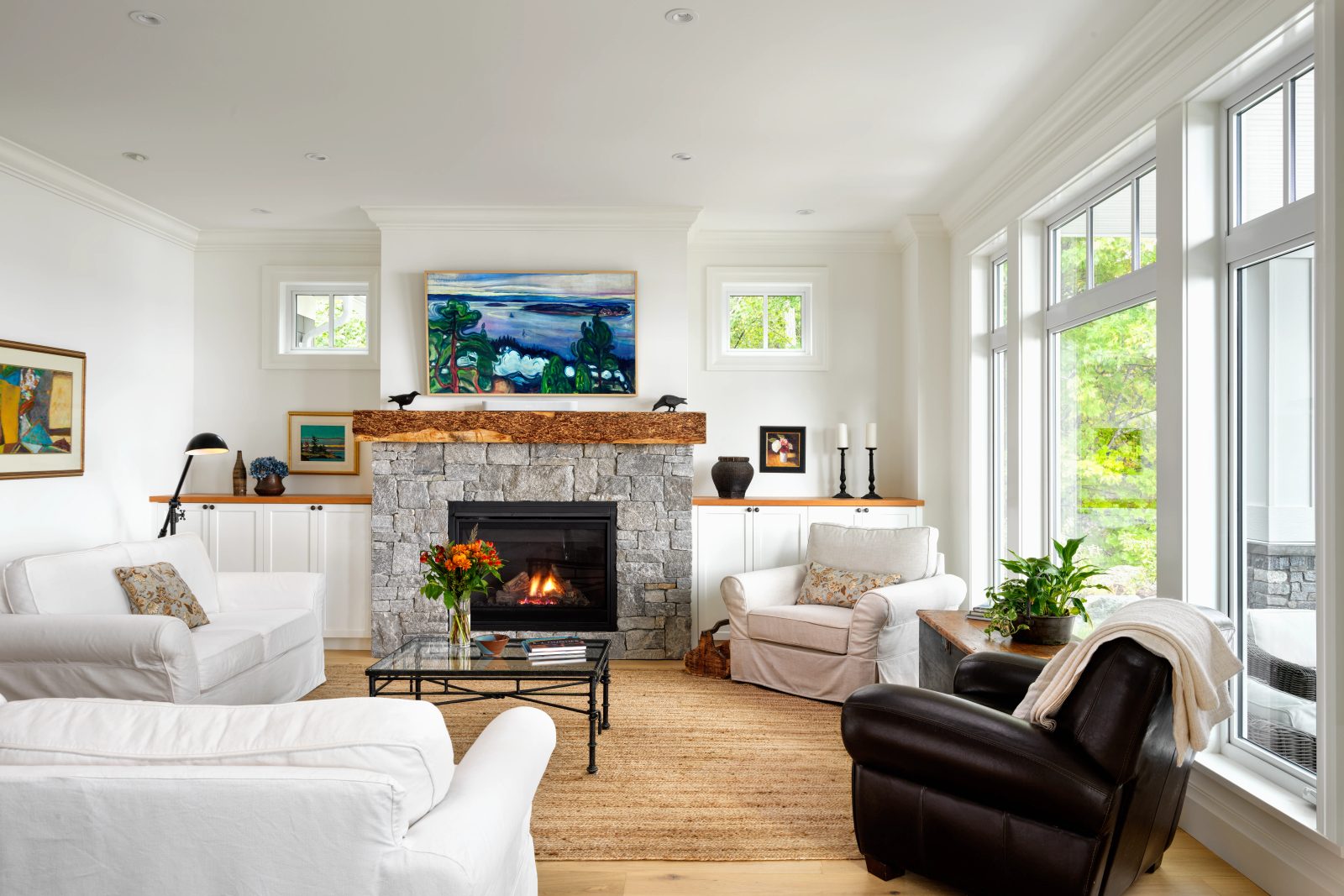 Living room with a grey stone fireplace under a live-edge timber mantel, white slipcovered chairs on a jute rug, and tall windows down one side.