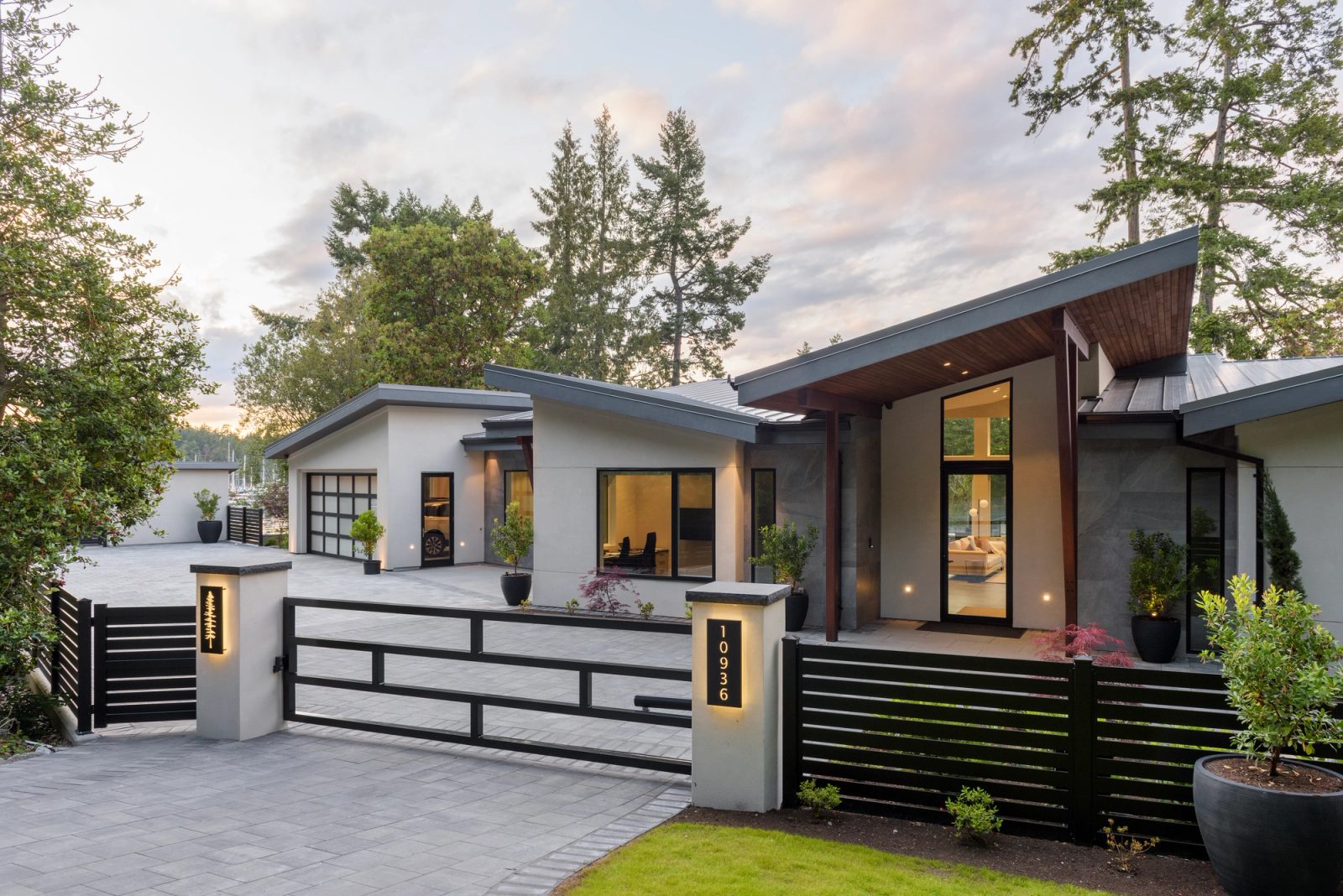 Gated entry court at dusk with a slatted metal fence, glazed garage door and a lit fir entry beyond
