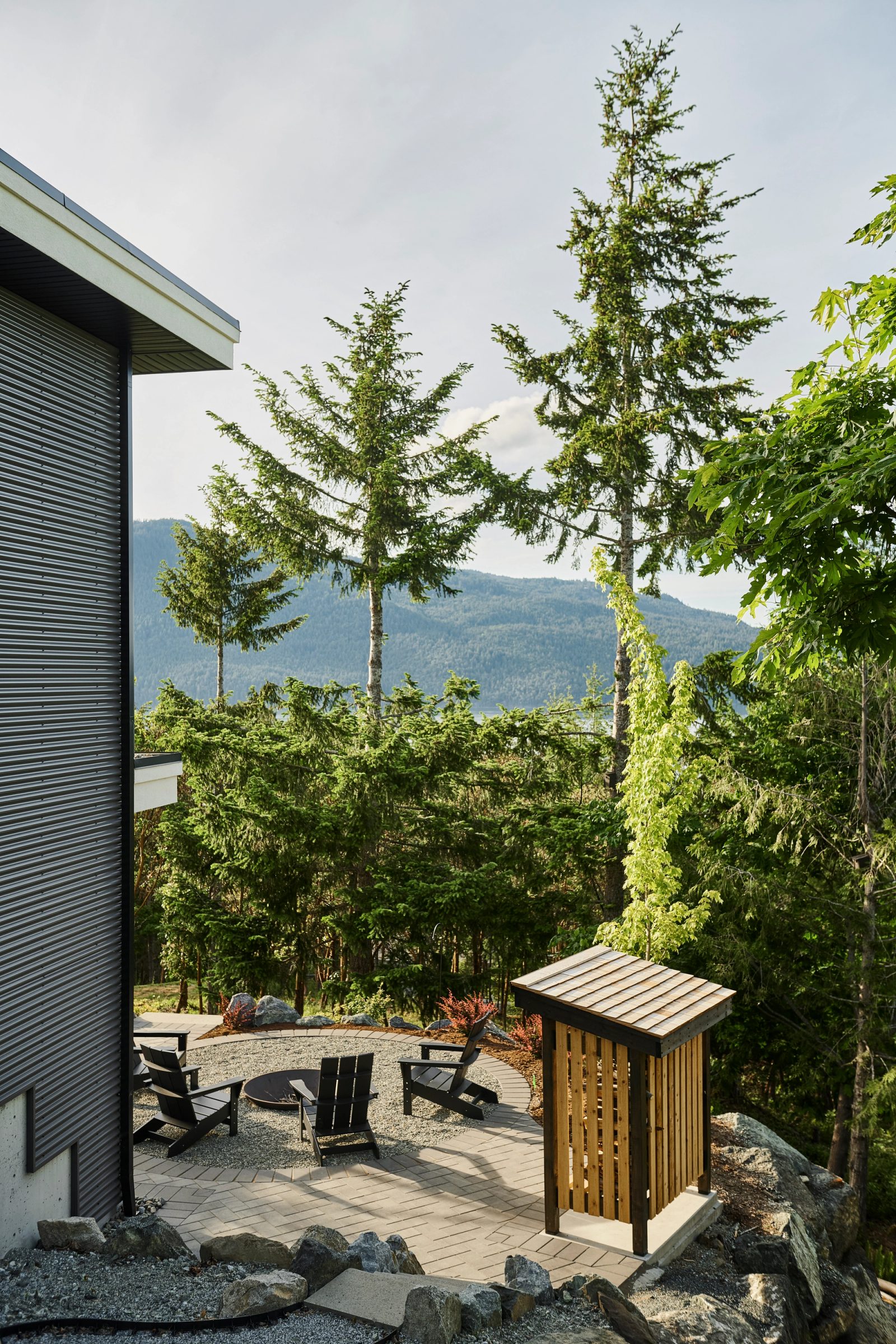 Fire pit terrace beside corrugated siding, firs and the far shore beyond the rock edge
