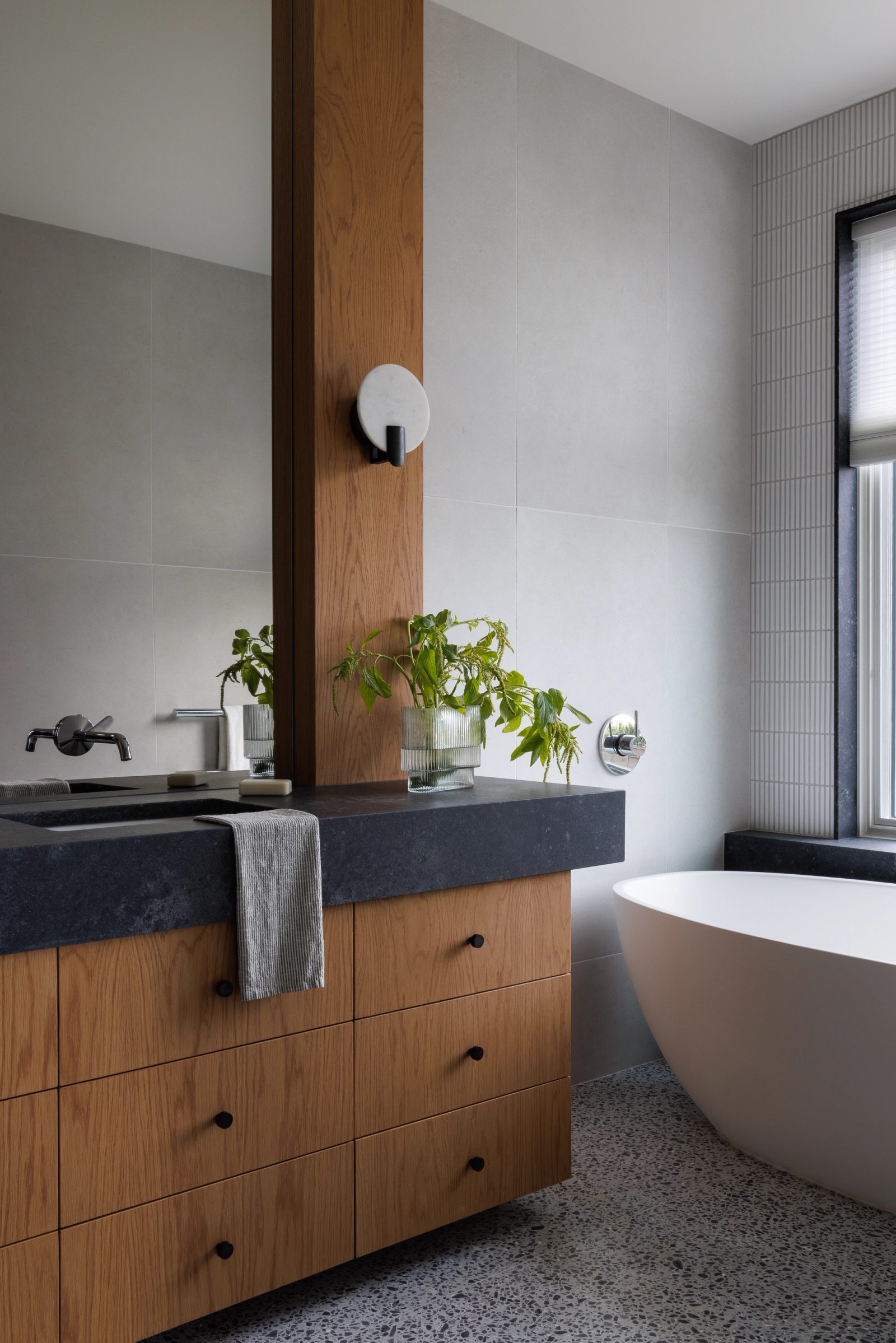 Left: a bathroom vanity in oak under dark stone, a tall mirror on an oak pier and a freestanding tub beyond.