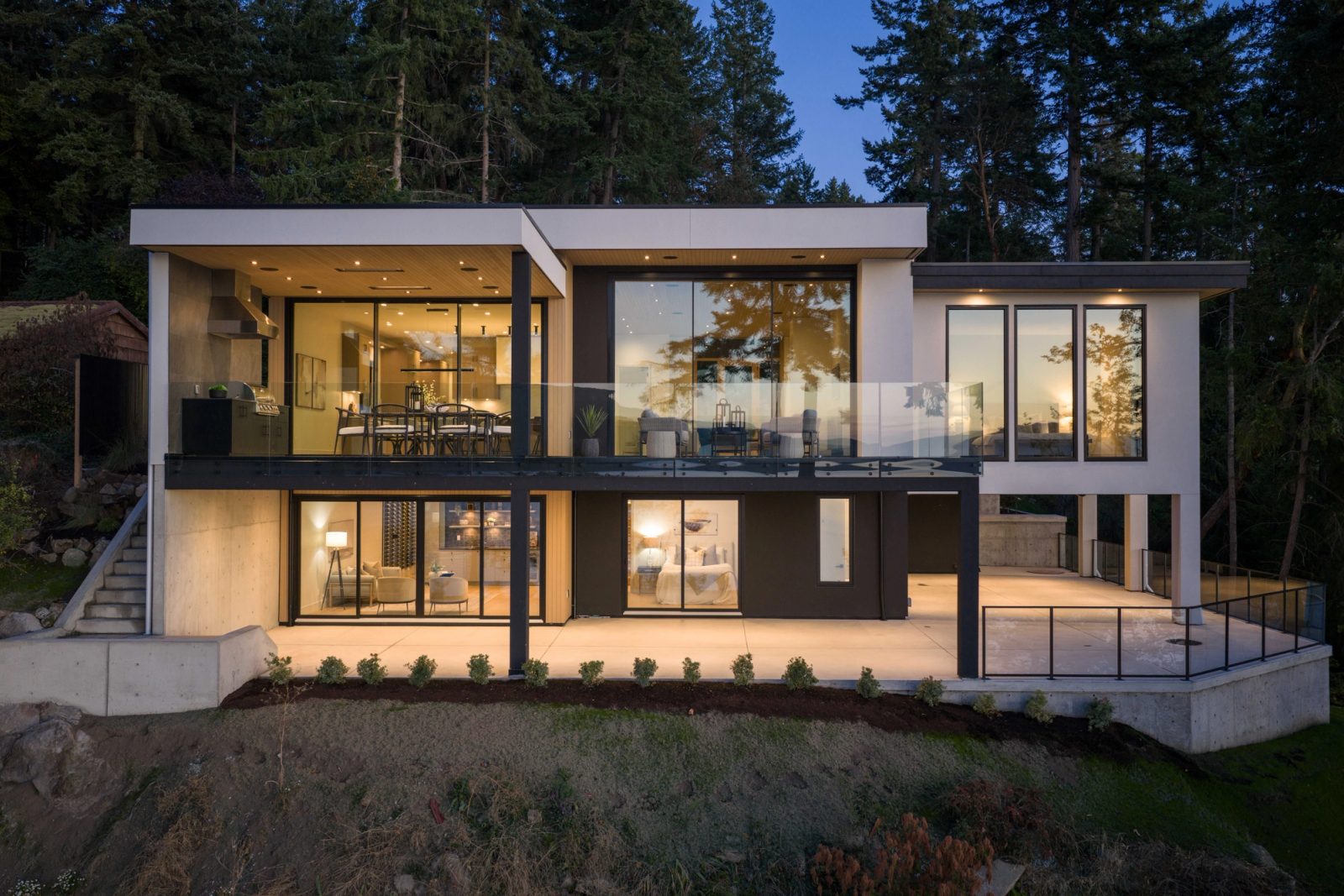 The whole house seen from below at dusk, both floors lit through full-height glass, standing on its concrete base among firs.