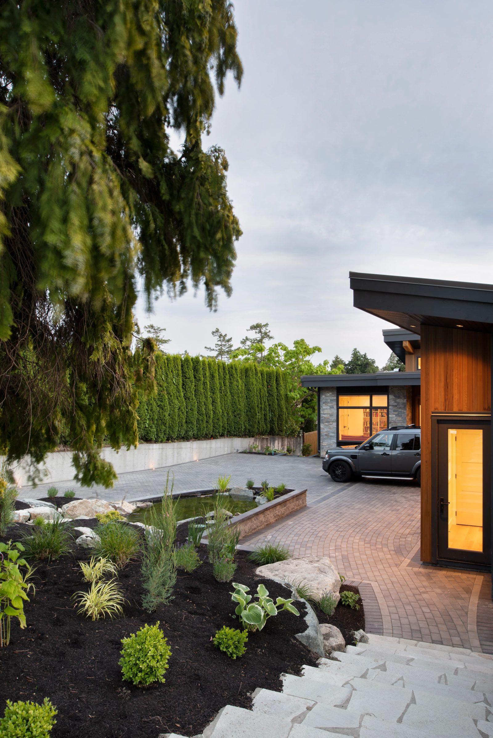 Paver driveway curving past a planted pond to a cedar-clad entry volume lit at dusk