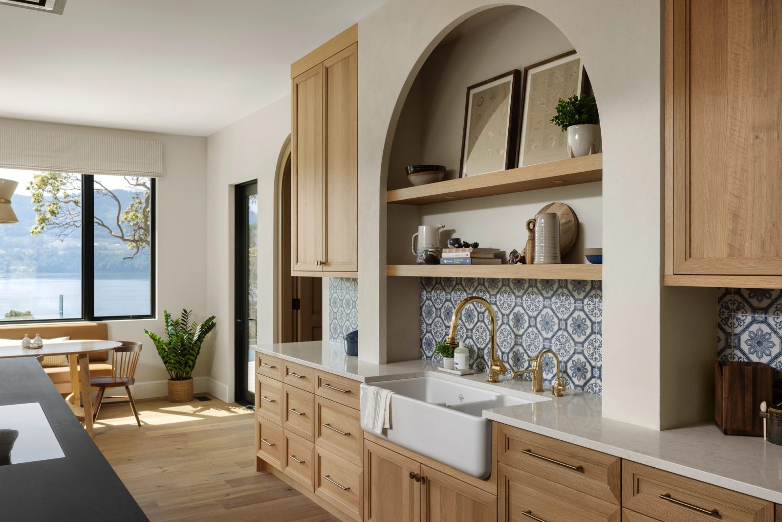 A kitchen with a farmhouse sink under an arched plaster niche and patterned blue tile, a banquette and the water framed in the window beyond.