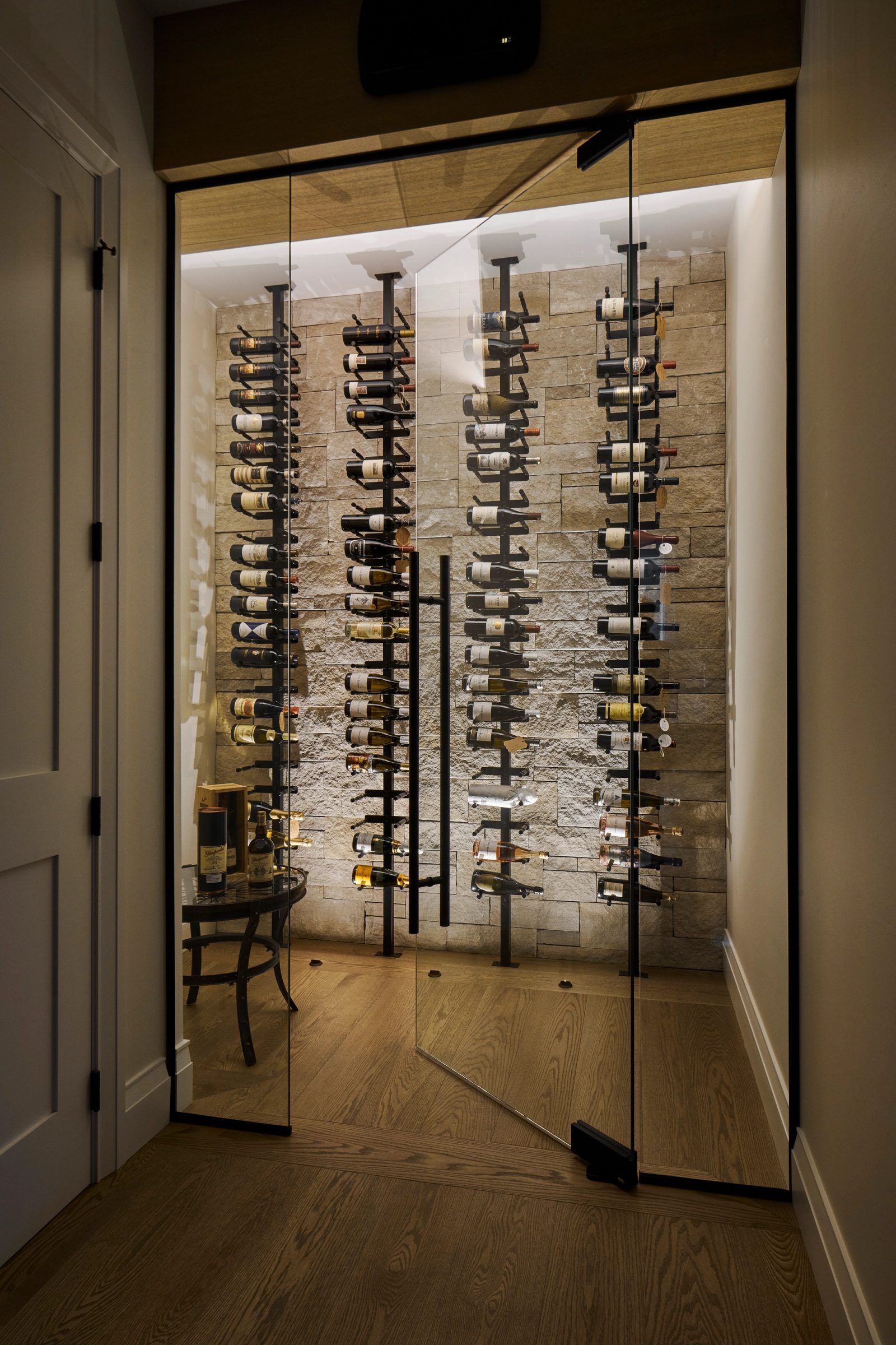 Wine wall in stacked stone behind frameless glass doors, bottles on black steel racks