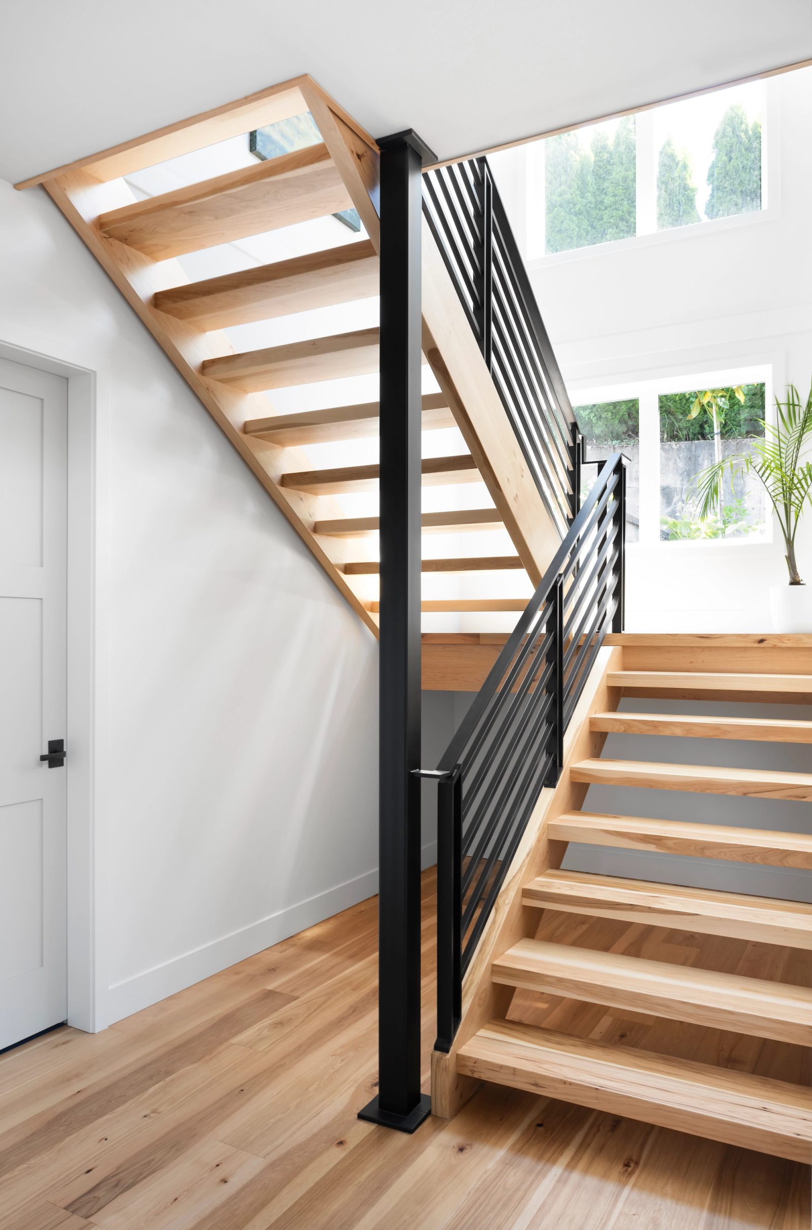 Open-riser hickory stair with a black horizontal steel rail turning at a landing below tall windows