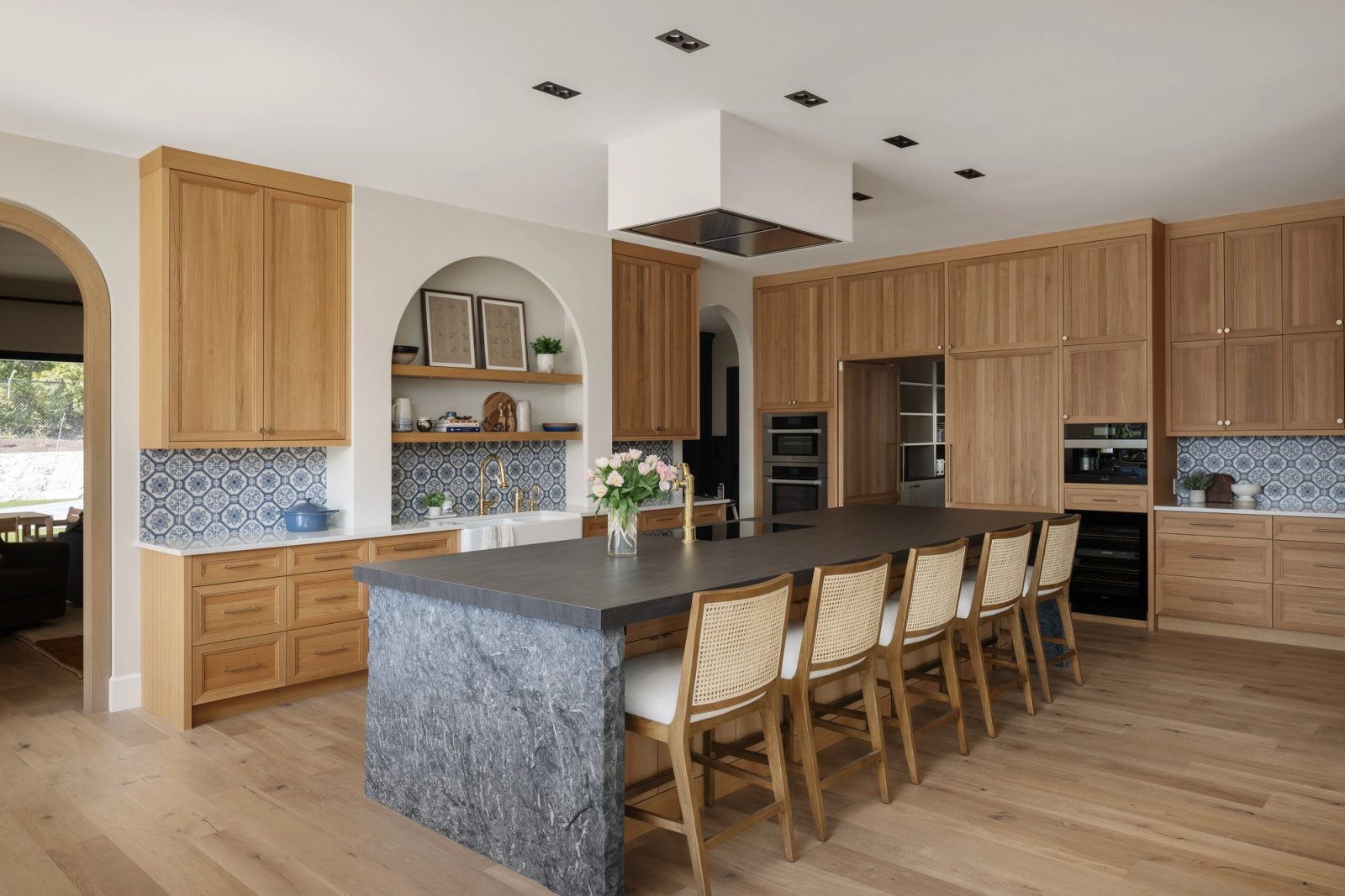 The length of a dark stone island with cane stools, oak cabinetry to the ceiling and the blue tile running behind.