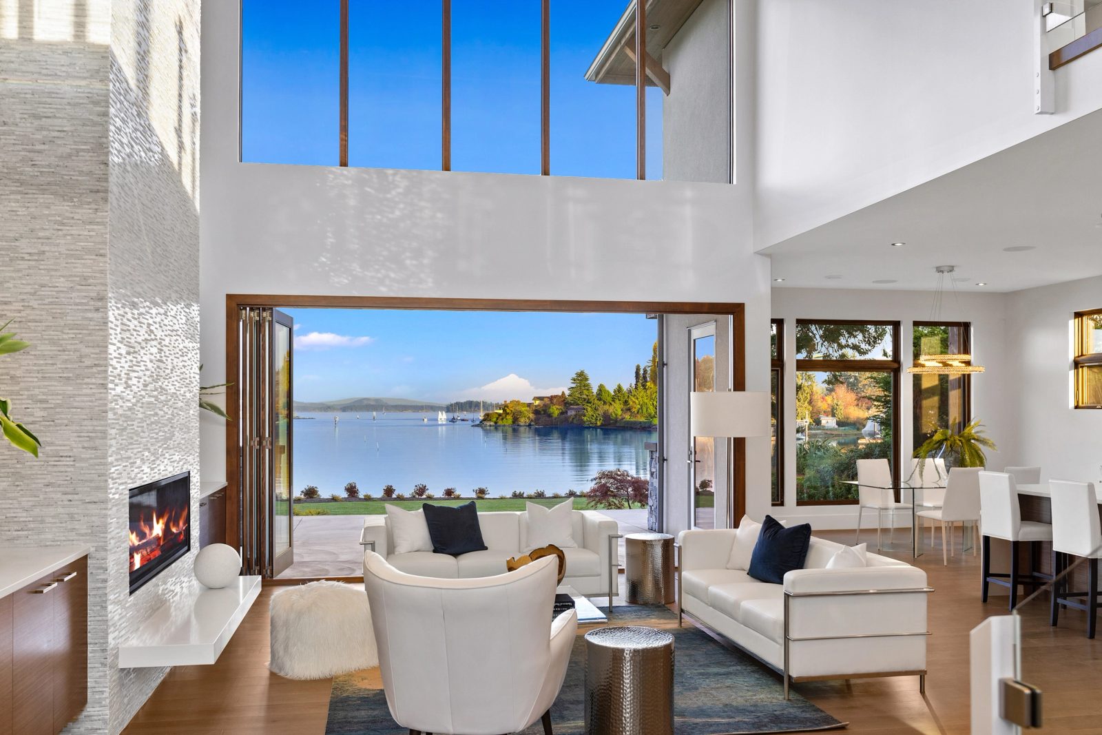 Two-storey living room with folding glass doors open to the harbour, a linear gas fireplace and a dining area beyond