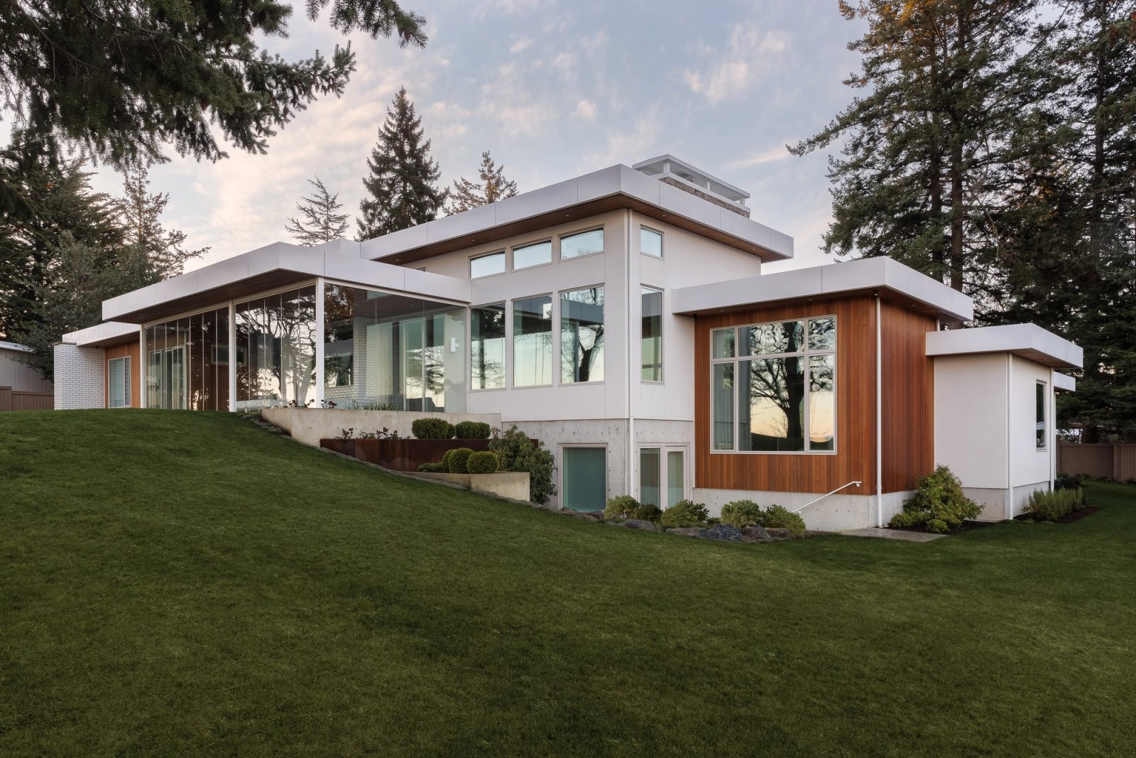 The seaward face of the house at dusk above a lawn falling away from it, a glazed corner returning to cedar, firs closing the frame on both sides.