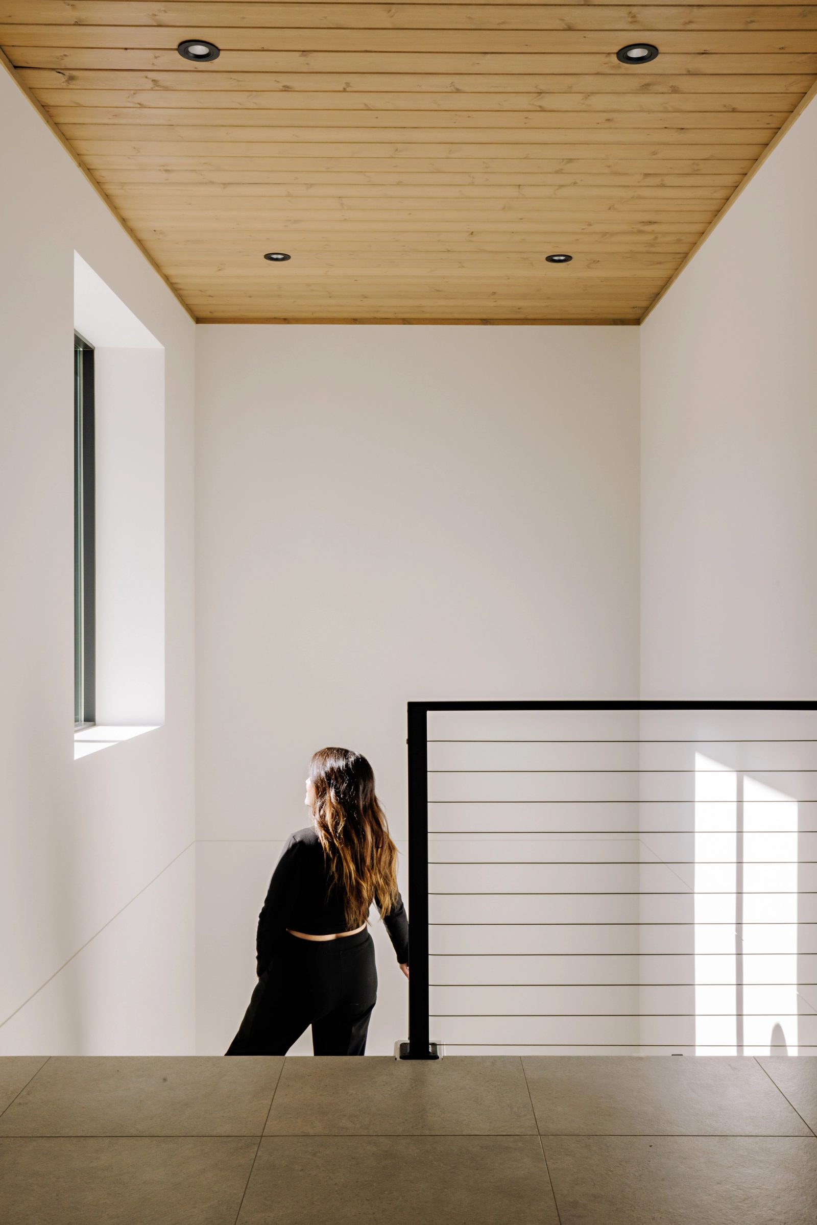 a tall white landing under a board ceiling, lit by a narrow vertical window, with a figure standing at a black cable rail.