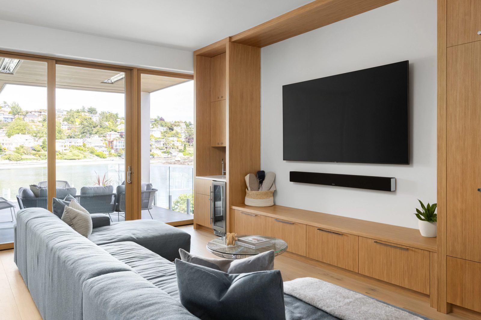 Family room with a wall-mounted screen on oak joinery, a deep sofa, and glass doors open to a terrace above the water.