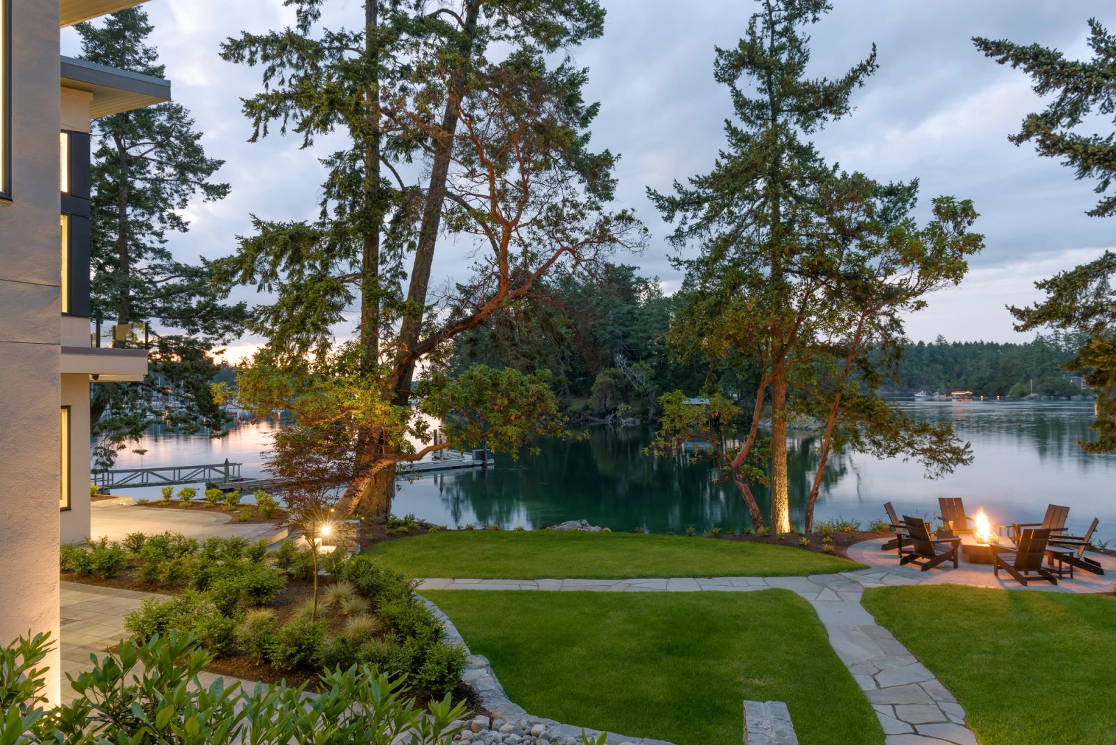Lawn and stone path leading to a firepit and dock at dusk, with the channel and forested shore beyond