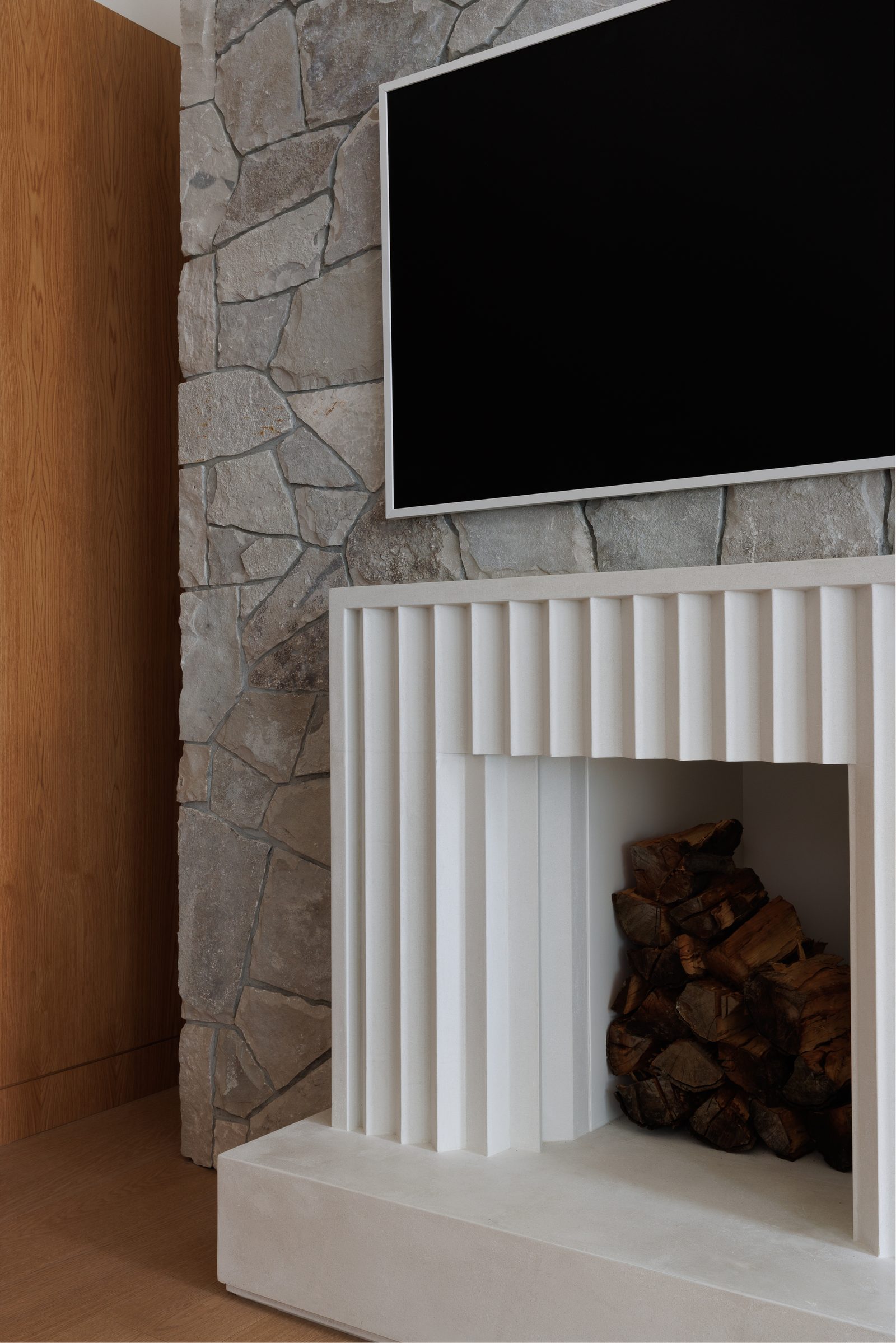 Right: the fireplace close, its fluted surround stepping out from rough stone coursing, split wood stacked in the opening.