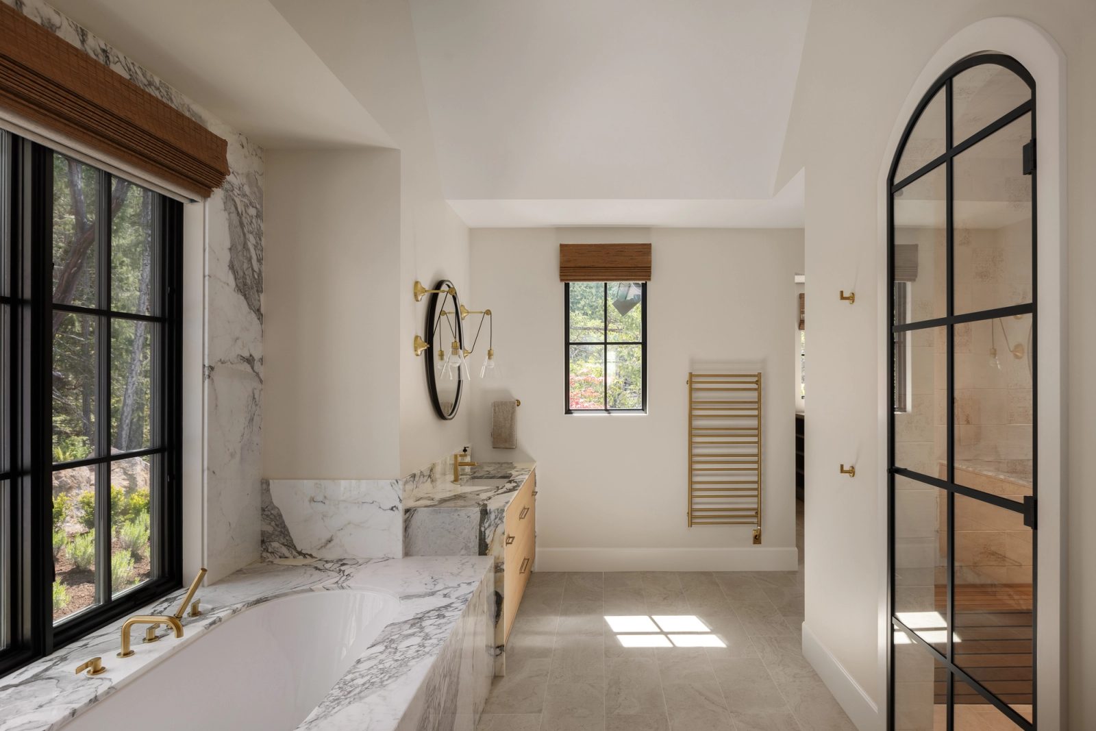 A bathroom with a marble-clad tub set into a black-framed window, a long marble counter opposite and an arched steel shower door.