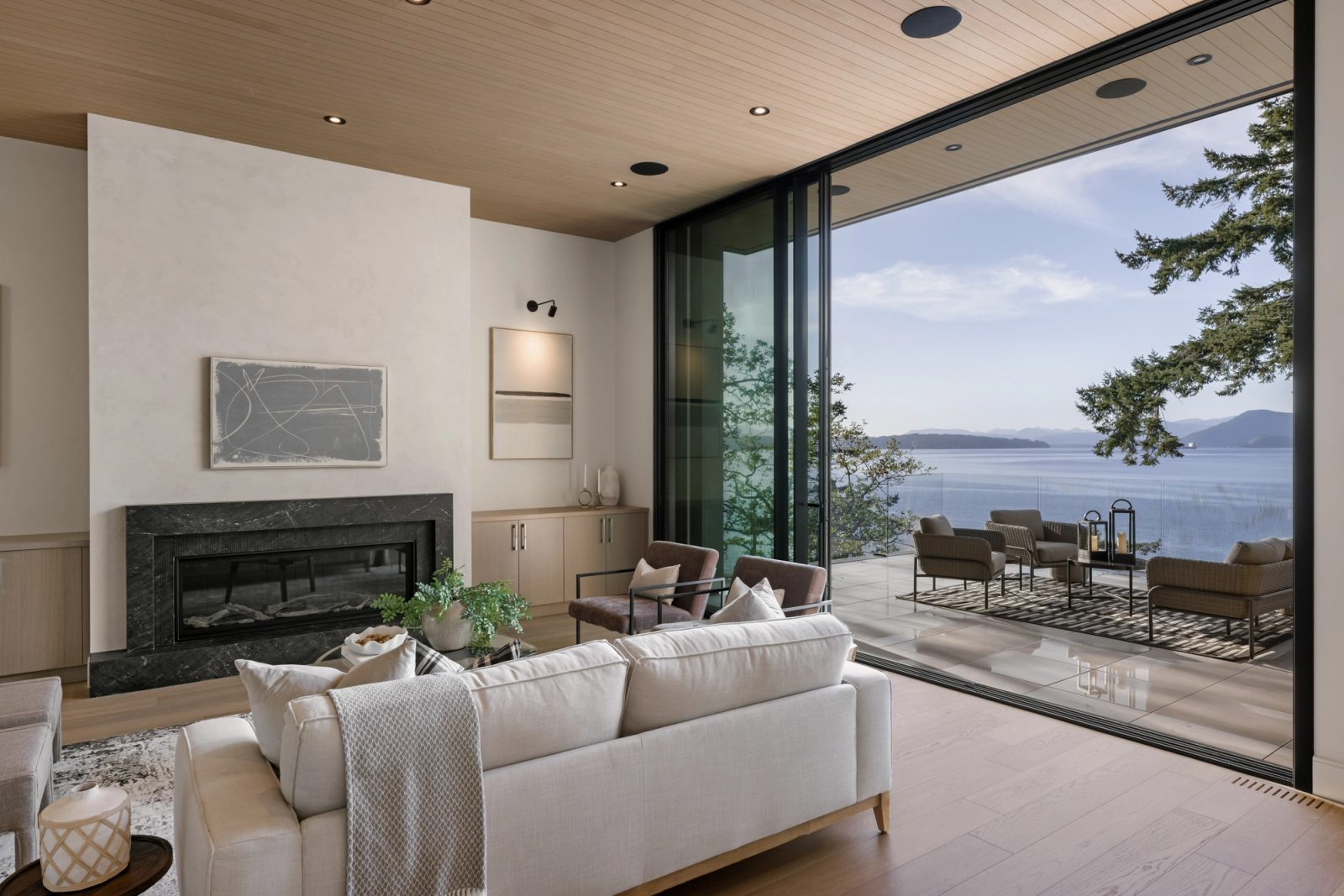 A living room with a plaster fireplace and dark stone hearth, one full wall slid open onto a tiled terrace and the water beyond.