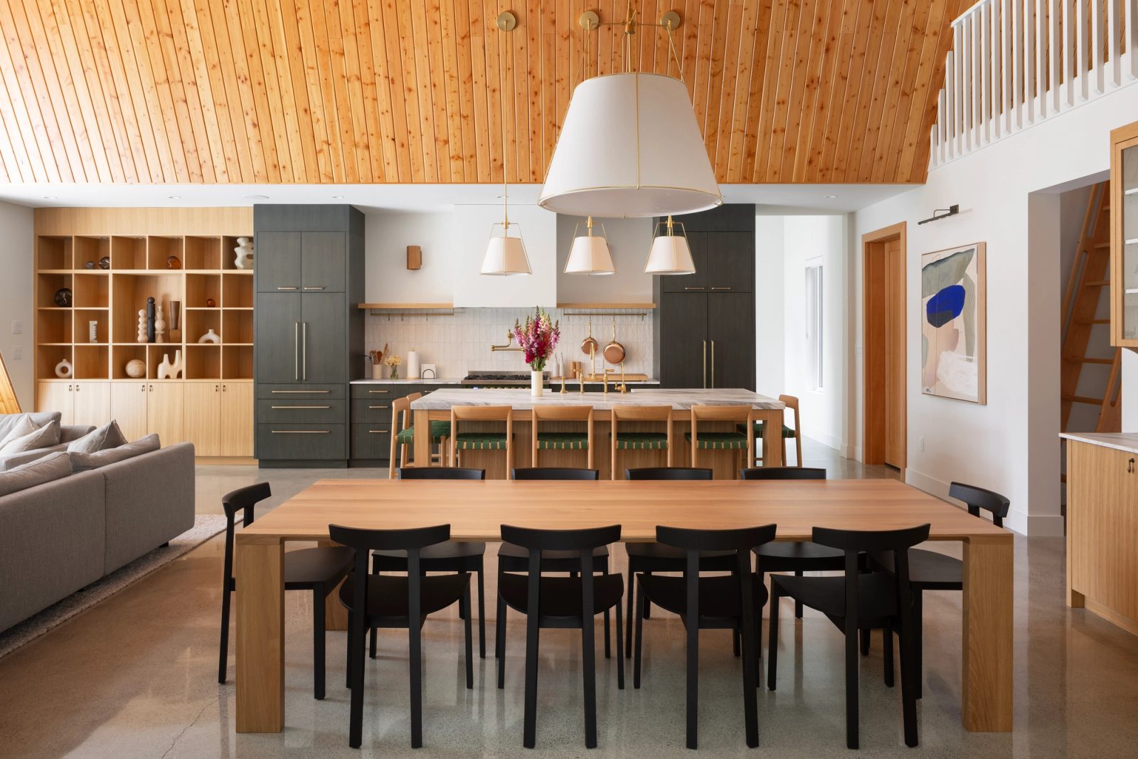 Long oak dining table facing a marble island and dark green kitchen under a cedar ceiling