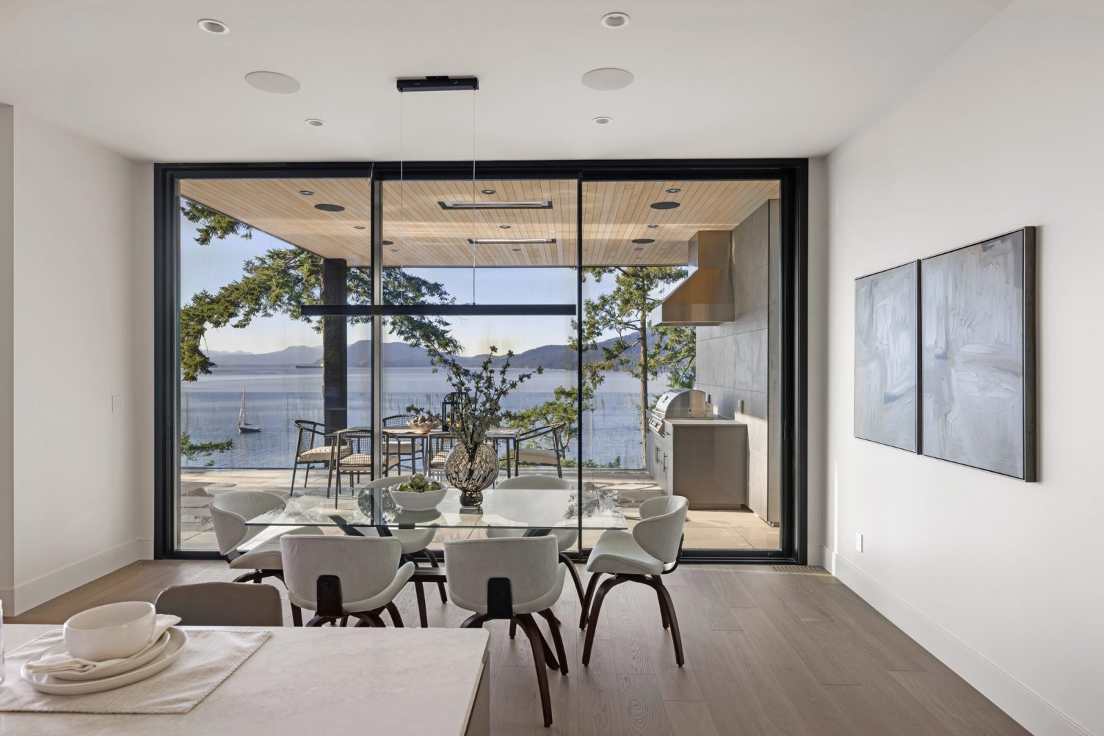 A dining table set in front of a sliding wall, opening to a covered terrace with an outdoor kitchen and a sailboat on the water past the rail.