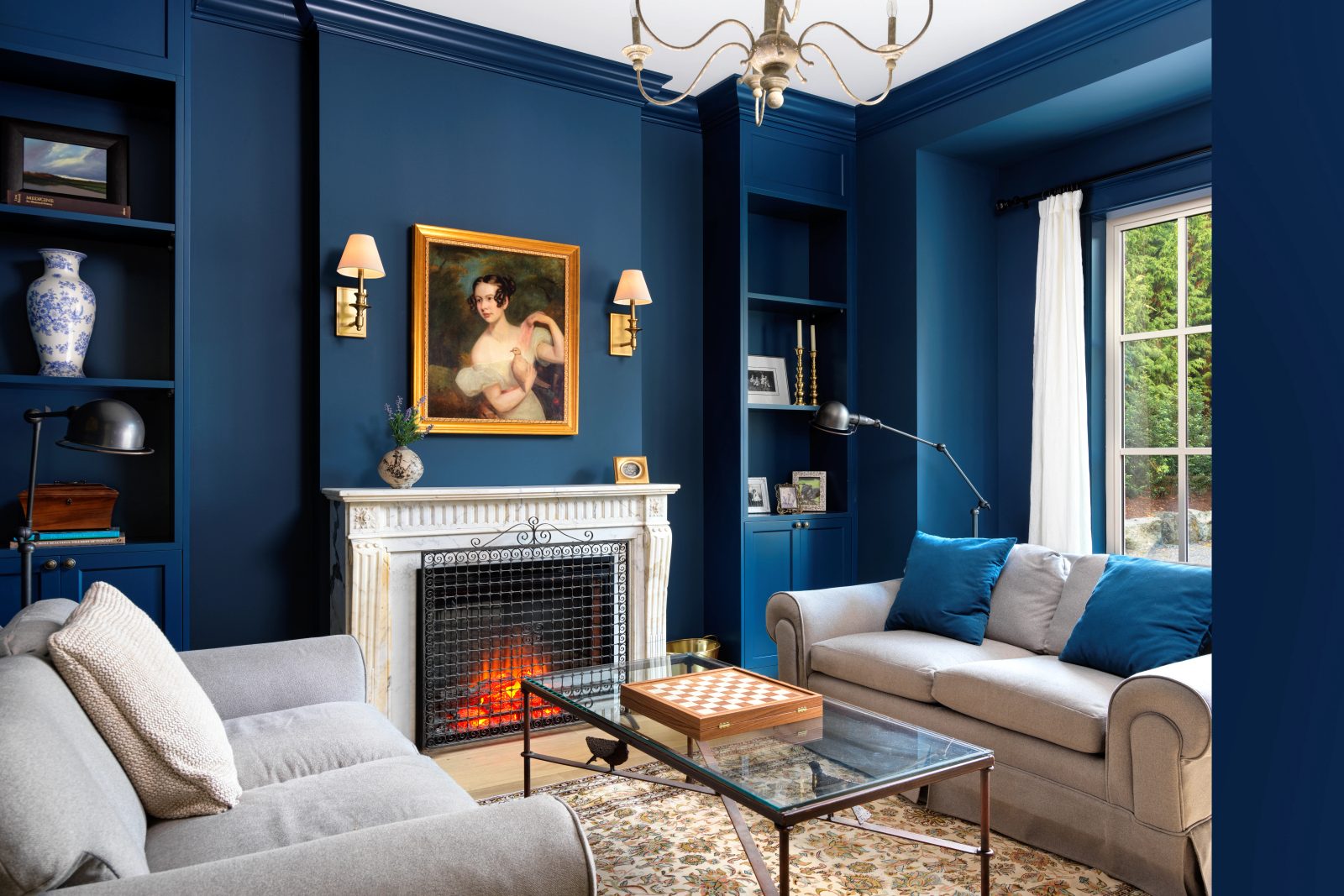 Small room painted deep blue to the ceiling, with built-in shelving, a carved white marble mantel and a portrait hung above it.