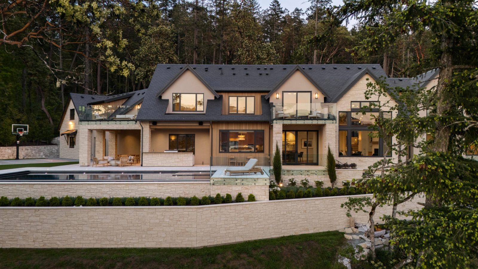 The house at dusk from across the pool, terraces and stone retaining walls stepping down to the water, forest closing behind.